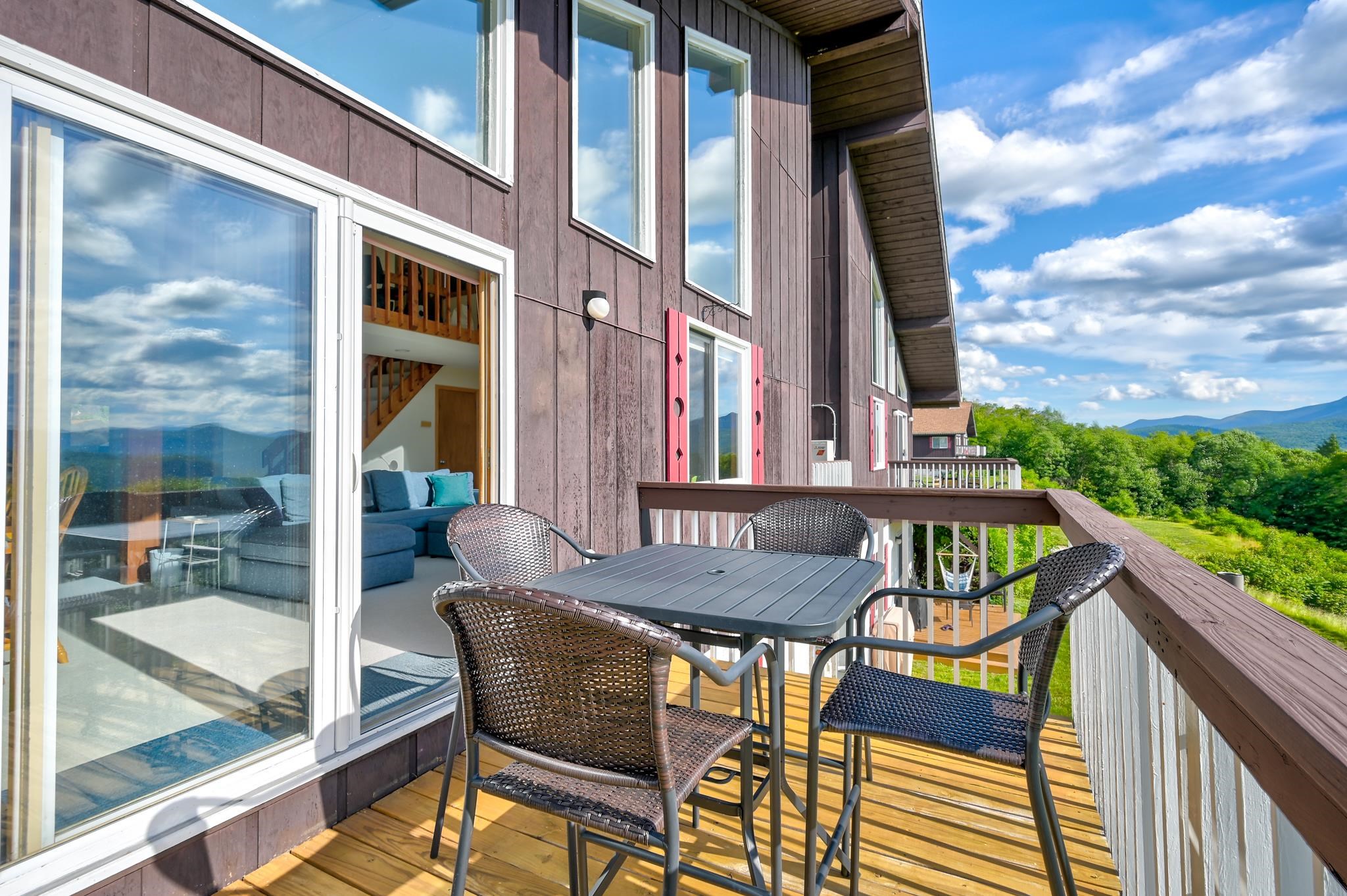 Furnished wooden deck with wicker chairs and a table overlooking green hills and distant mountains.