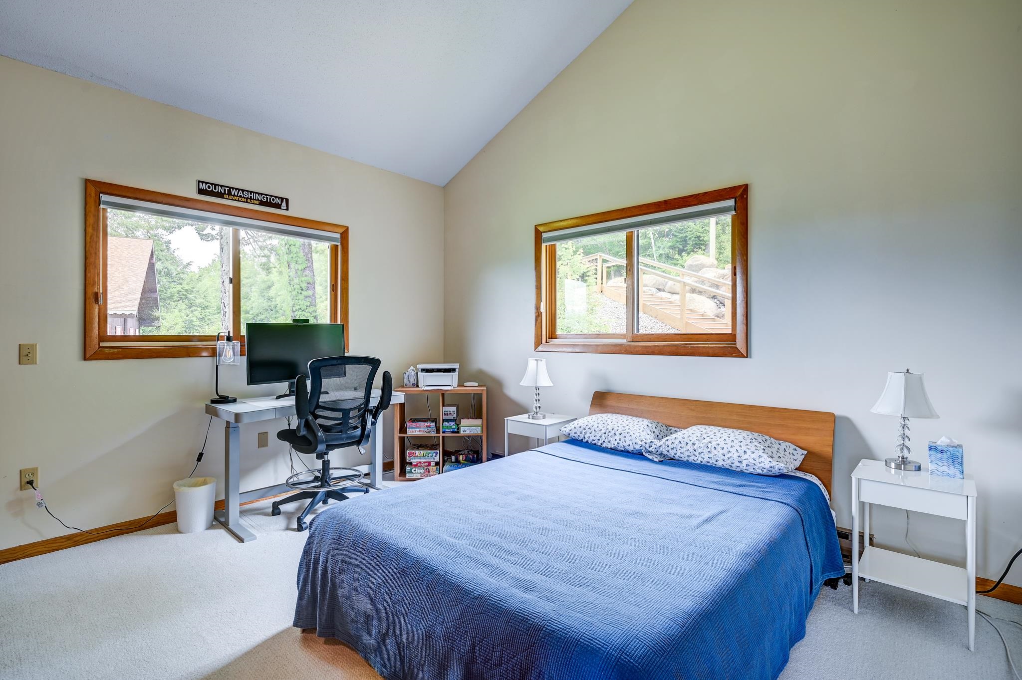 Bedroom with a blue-covered bed, vaulted ceiling, and desk beside two wood-trimmed windows.