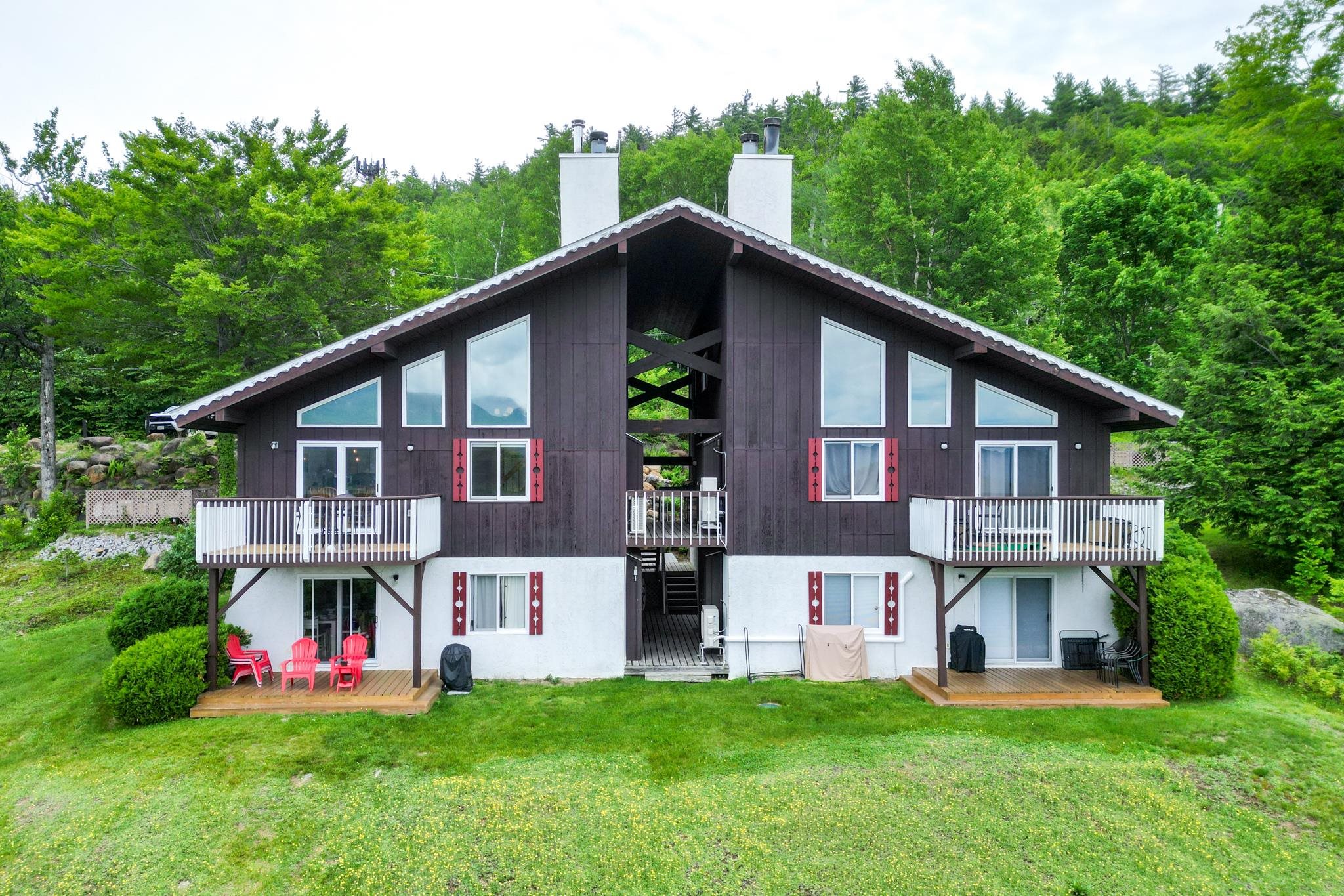 Chalet-style home with two balconies and red shutters, set against a wooded backdrop.