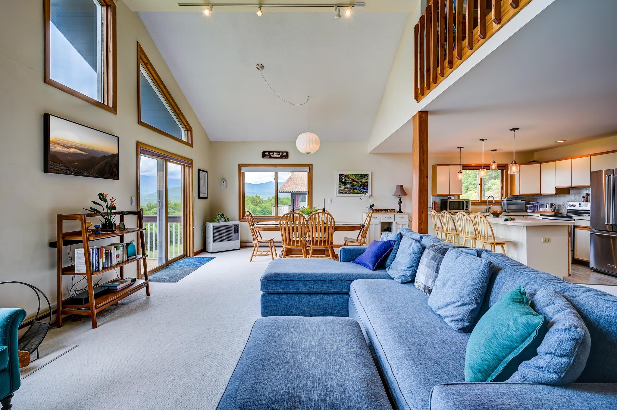 Open-plan living room with a blue sectional, dining area, kitchen, vaulted ceiling, and mountain views.