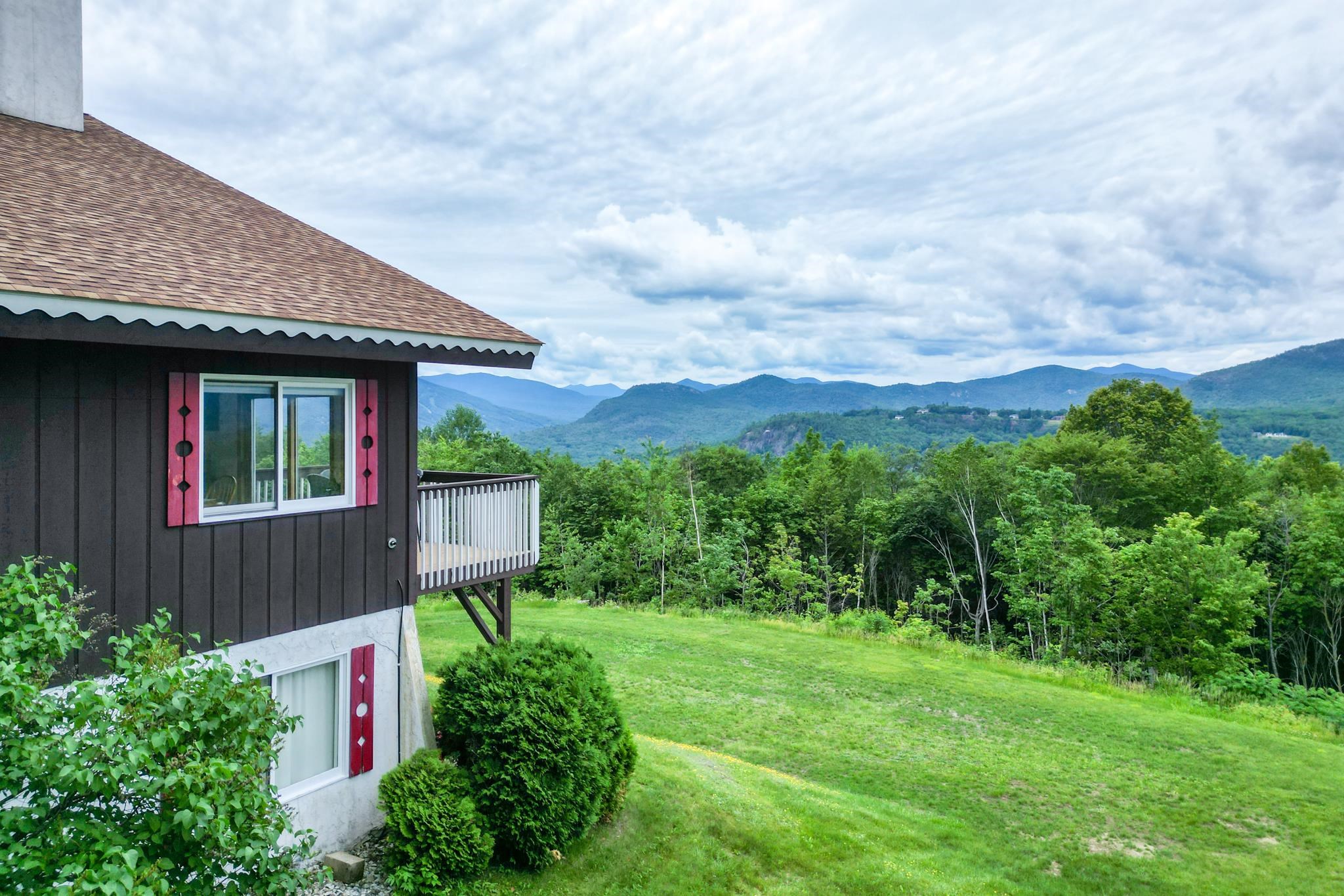 House with a small deck overlooking a green lawn, forest, and mountain ridges.
