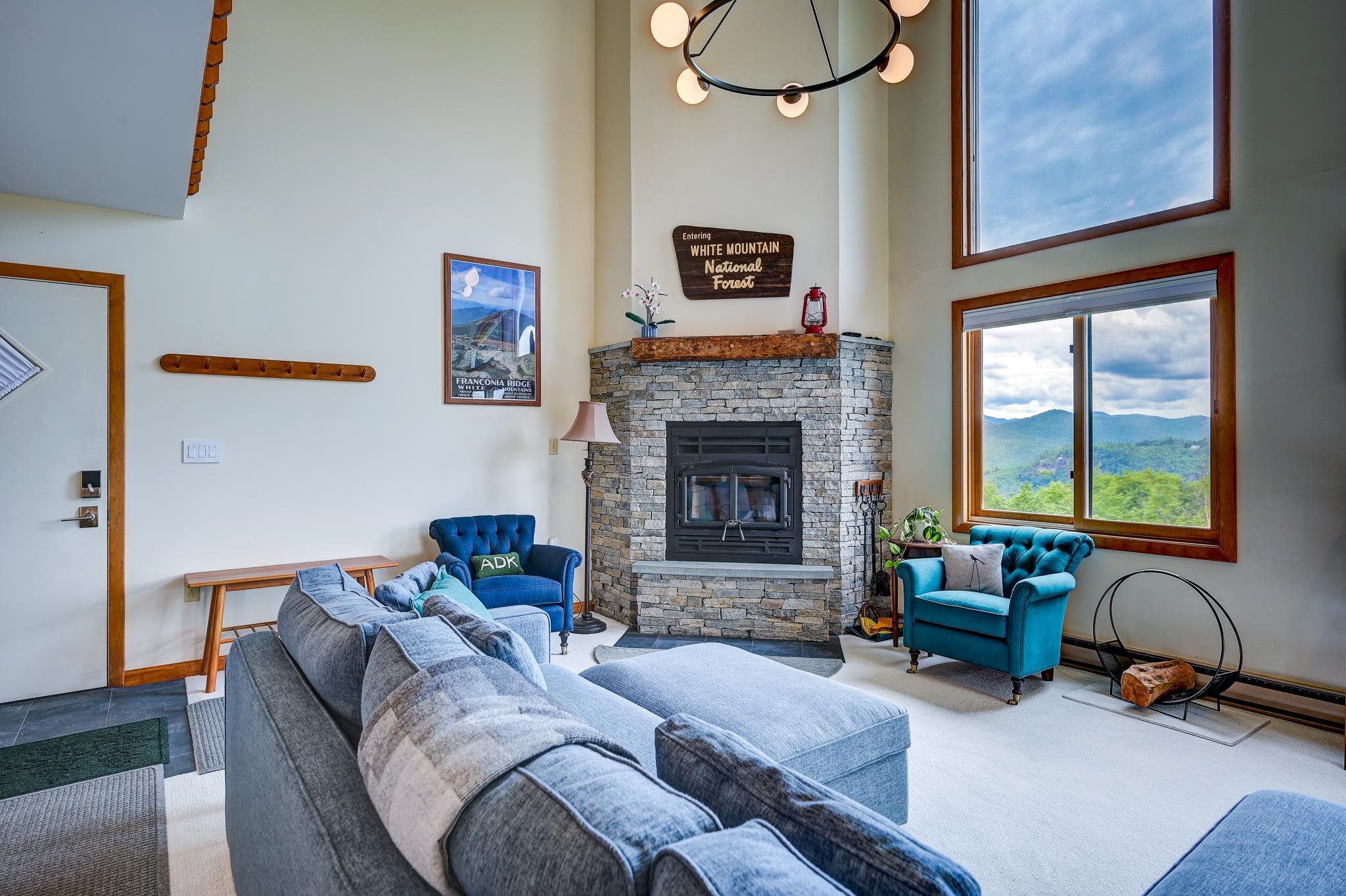 Living room with a stone fireplace, blue chairs, and large windows overlooking forested mountains.