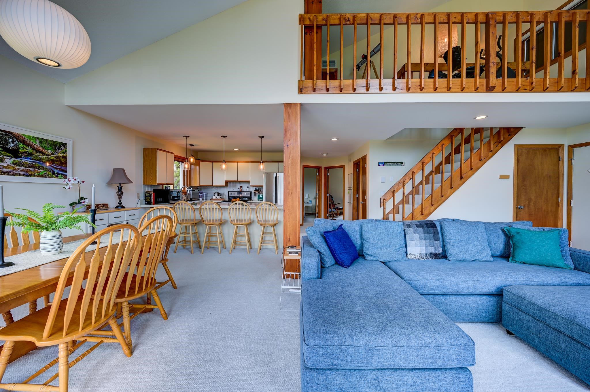 Open living area with a blue sectional, wooden dining set, kitchen island, and staircase to a loft.