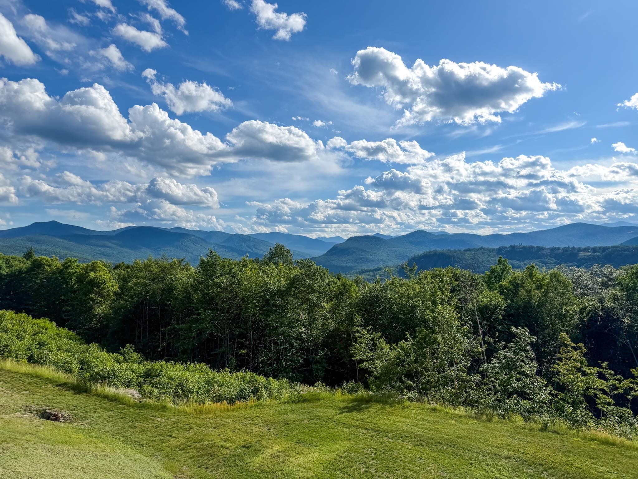 Mountain views beyond a grassy lawn and wooded hills beneath a cloud-filled blue sky.
