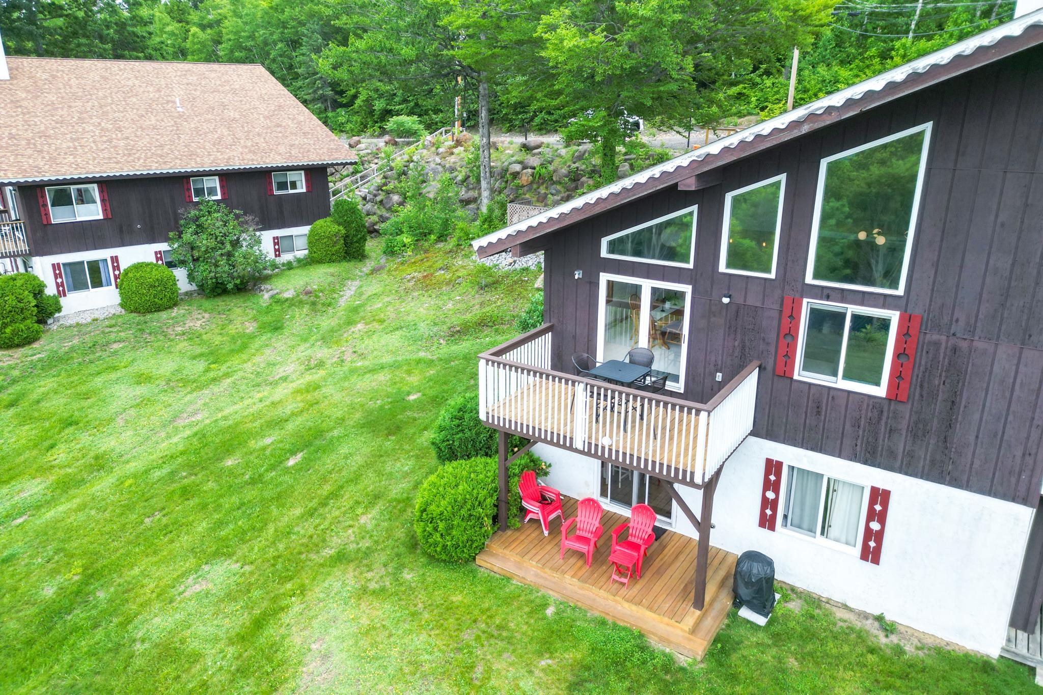 Dark-sided house with red shutters, a raised balcony, and a wooden deck with red chairs.