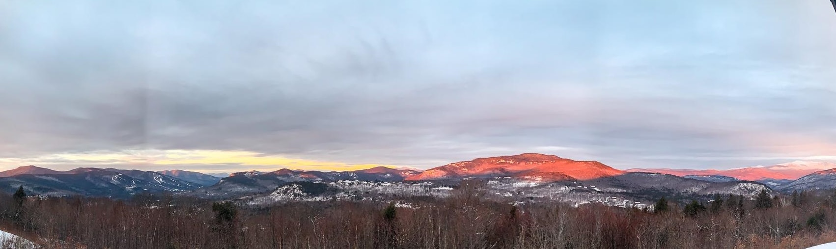 Snow-dusted mountain panorama glowing in the sunset above leafless woodland.