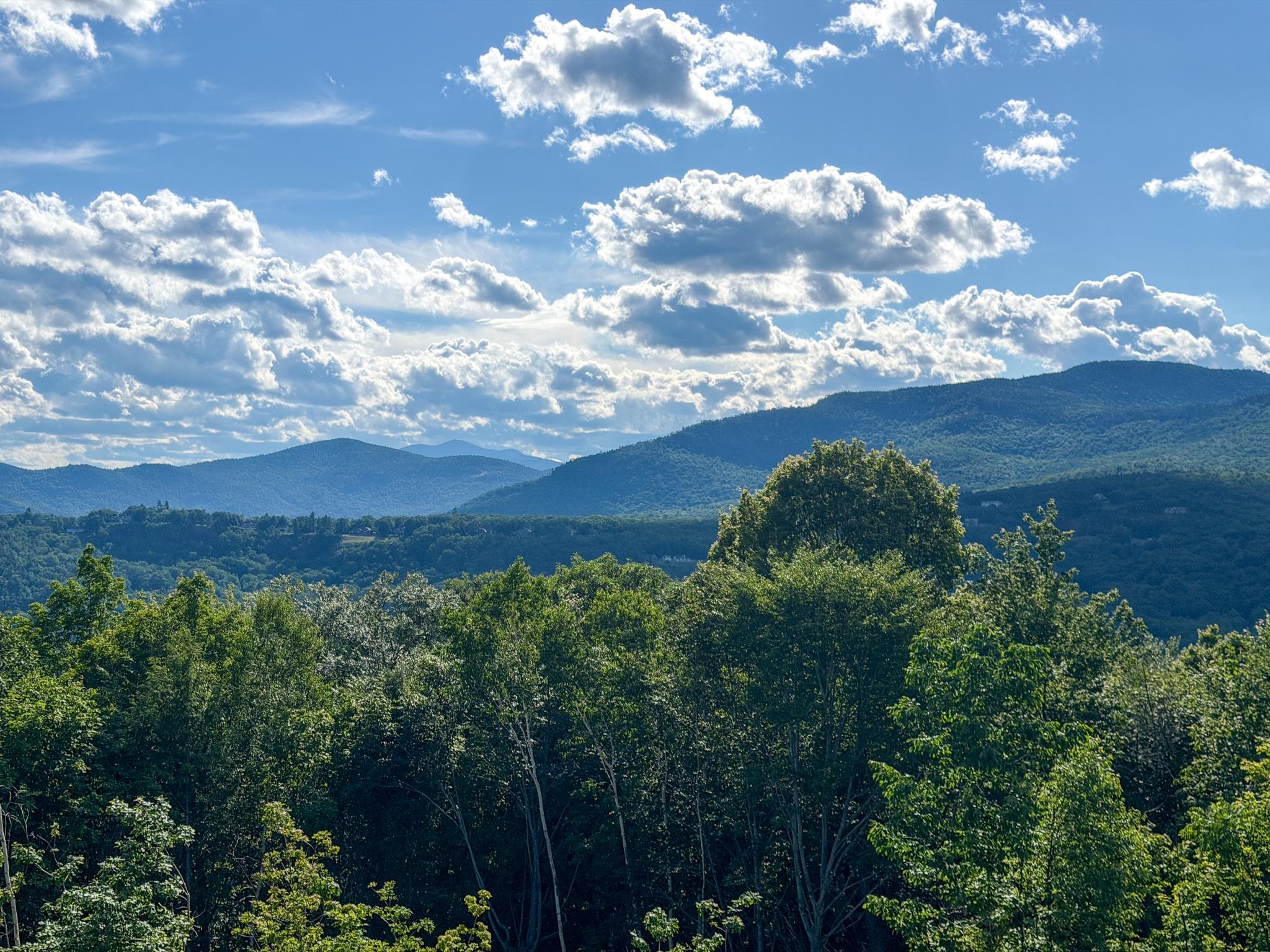 Mountain view across wooded hills beneath a cloud-filled blue sky.