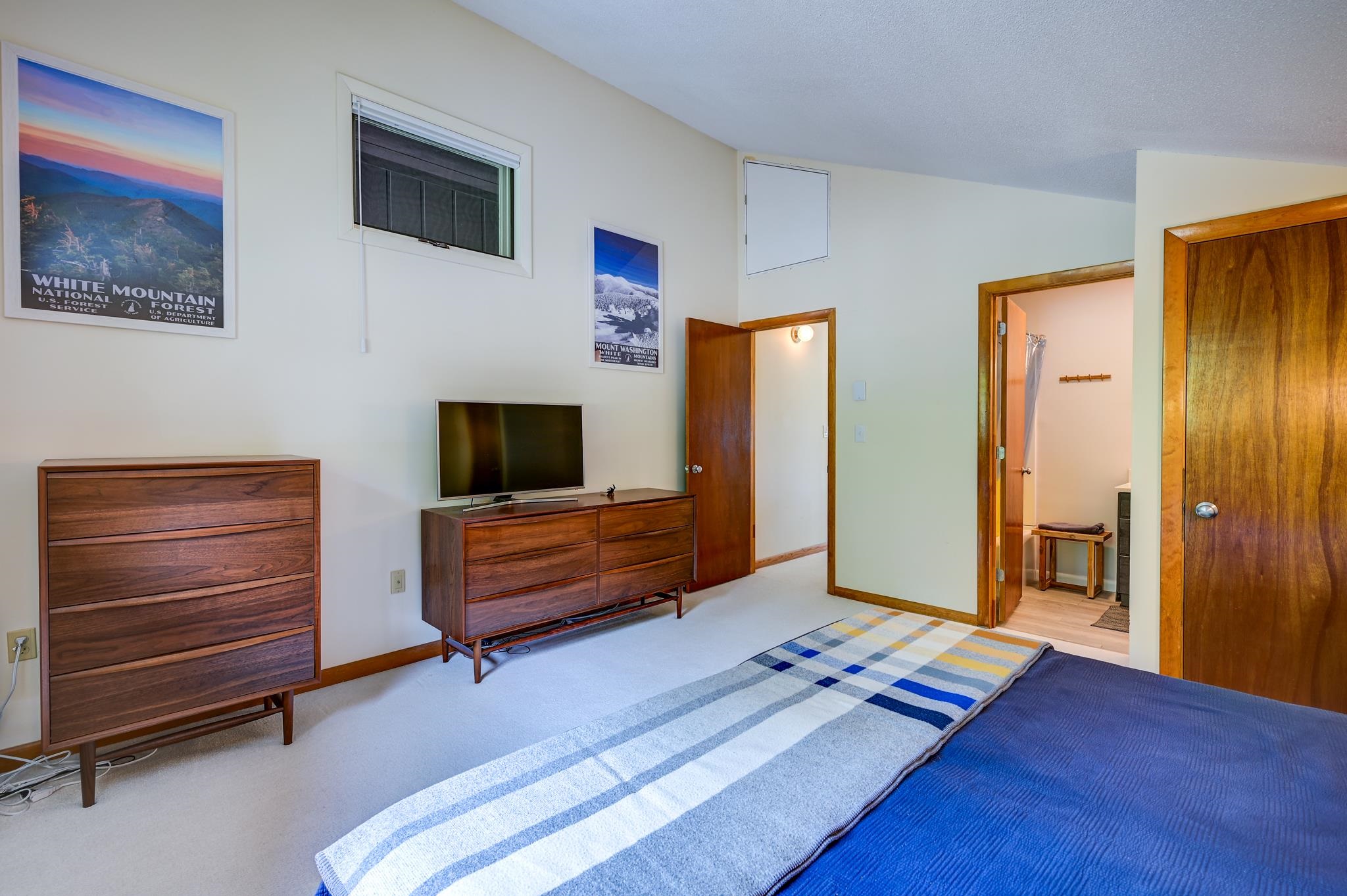 Furnished bedroom with a blue bedspread, wooden dressers, television, and White Mountain posters.