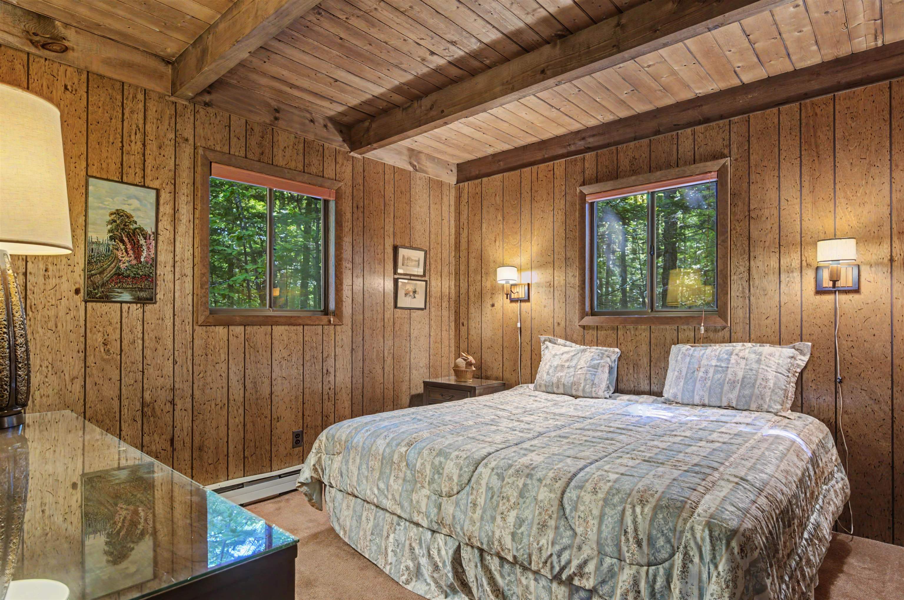 Bedroom with wood-paneled walls, exposed ceiling beams, and windows overlooking trees.
