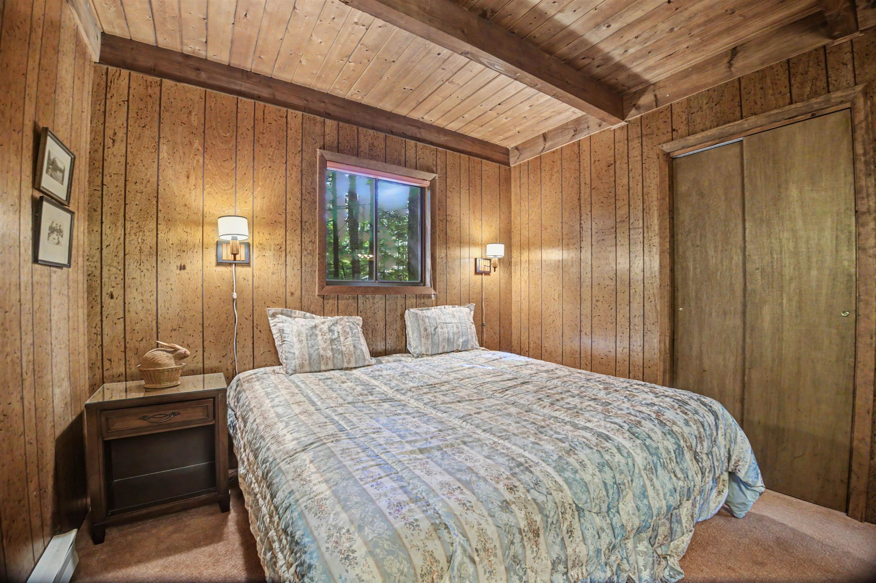 Furnished bedroom with wood-paneled walls, exposed ceiling beams, and a large bed beneath a window.