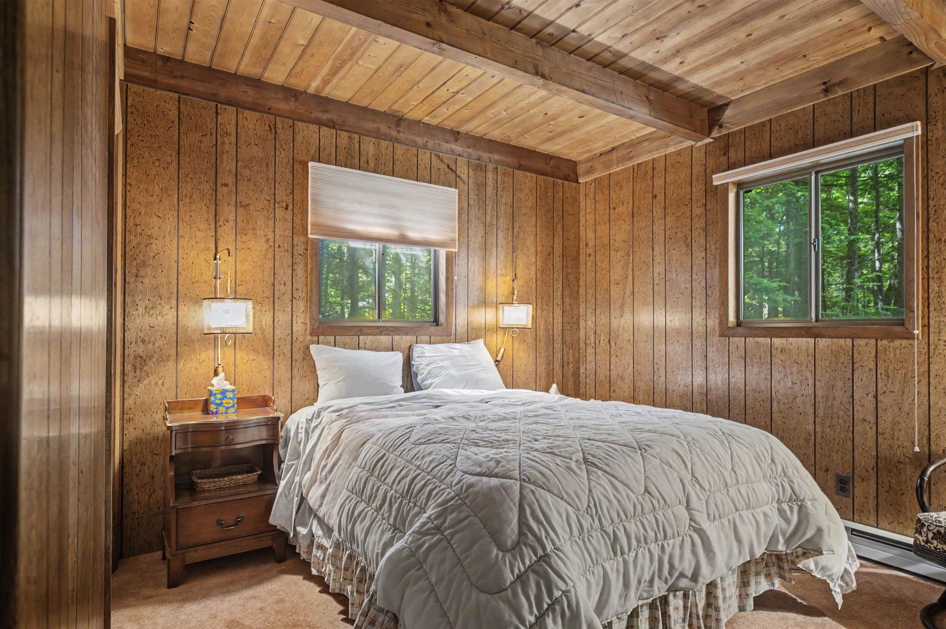 Bedroom with wood-paneled walls, vaulted beamed ceiling, and windows overlooking trees.