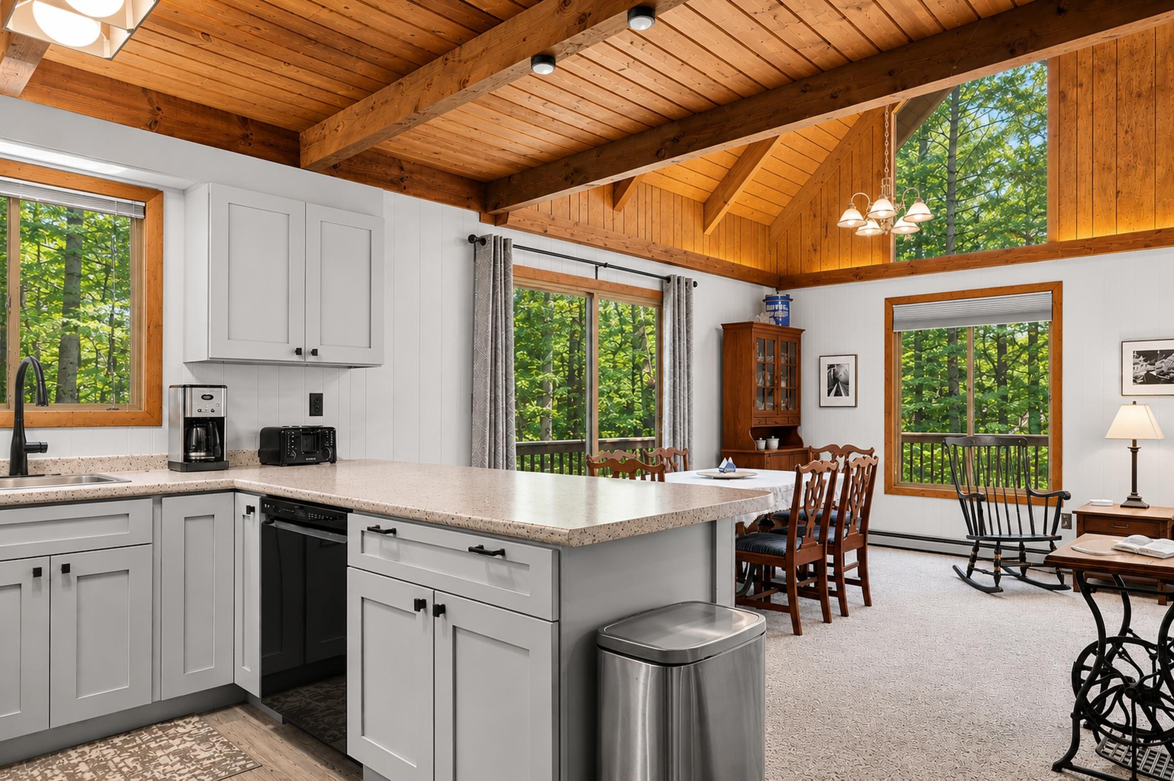 Open kitchen and dining area with a white island, vaulted wood ceiling, and windows overlooking leafy trees.