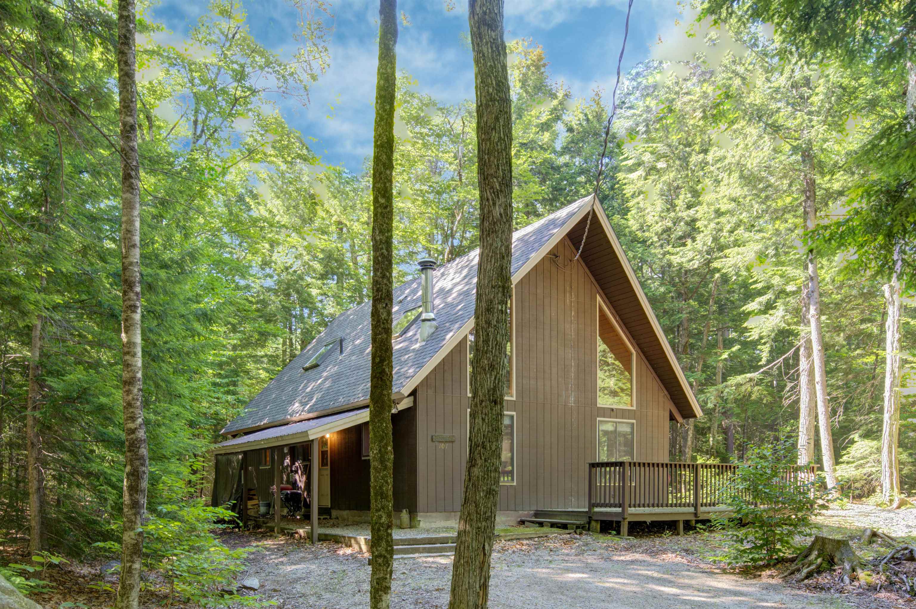 Brown A-frame house with a steep shingle roof and small deck among leafy trees.