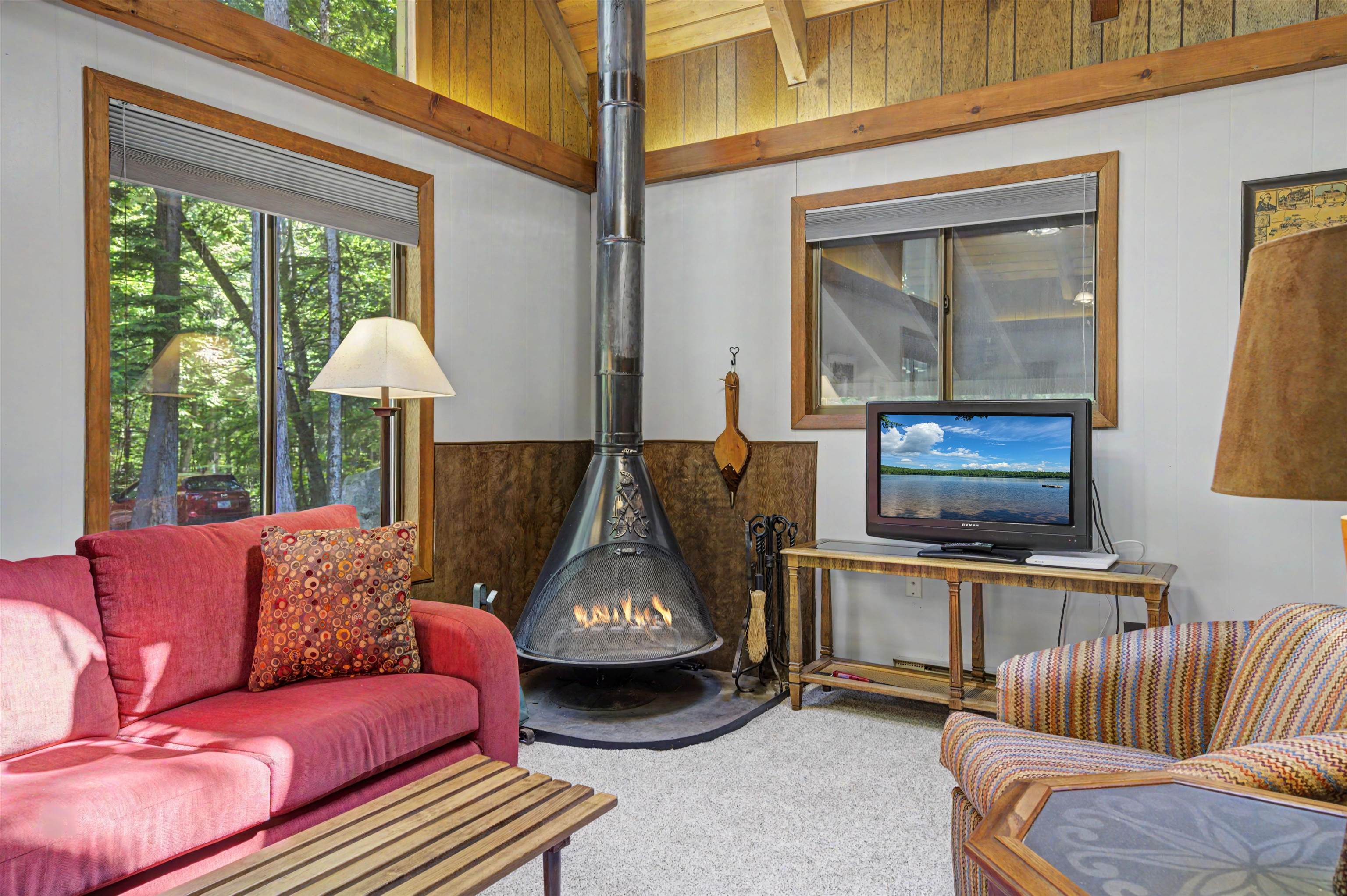 Living room with a freestanding fireplace, red sofa, and large windows overlooking trees.