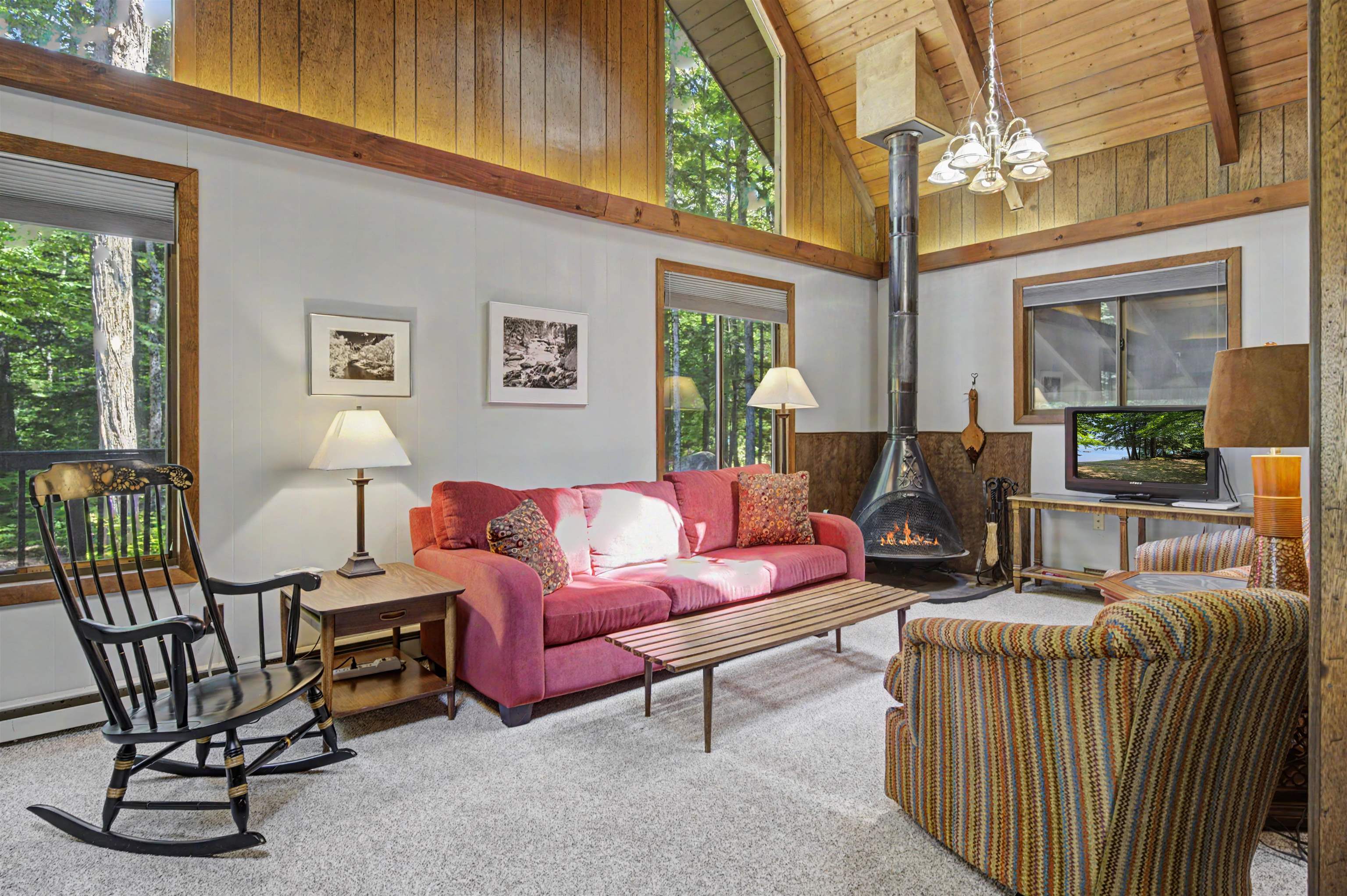 Living room with vaulted wood ceiling, pink sofa, large wooded-view windows, and freestanding fireplace.
