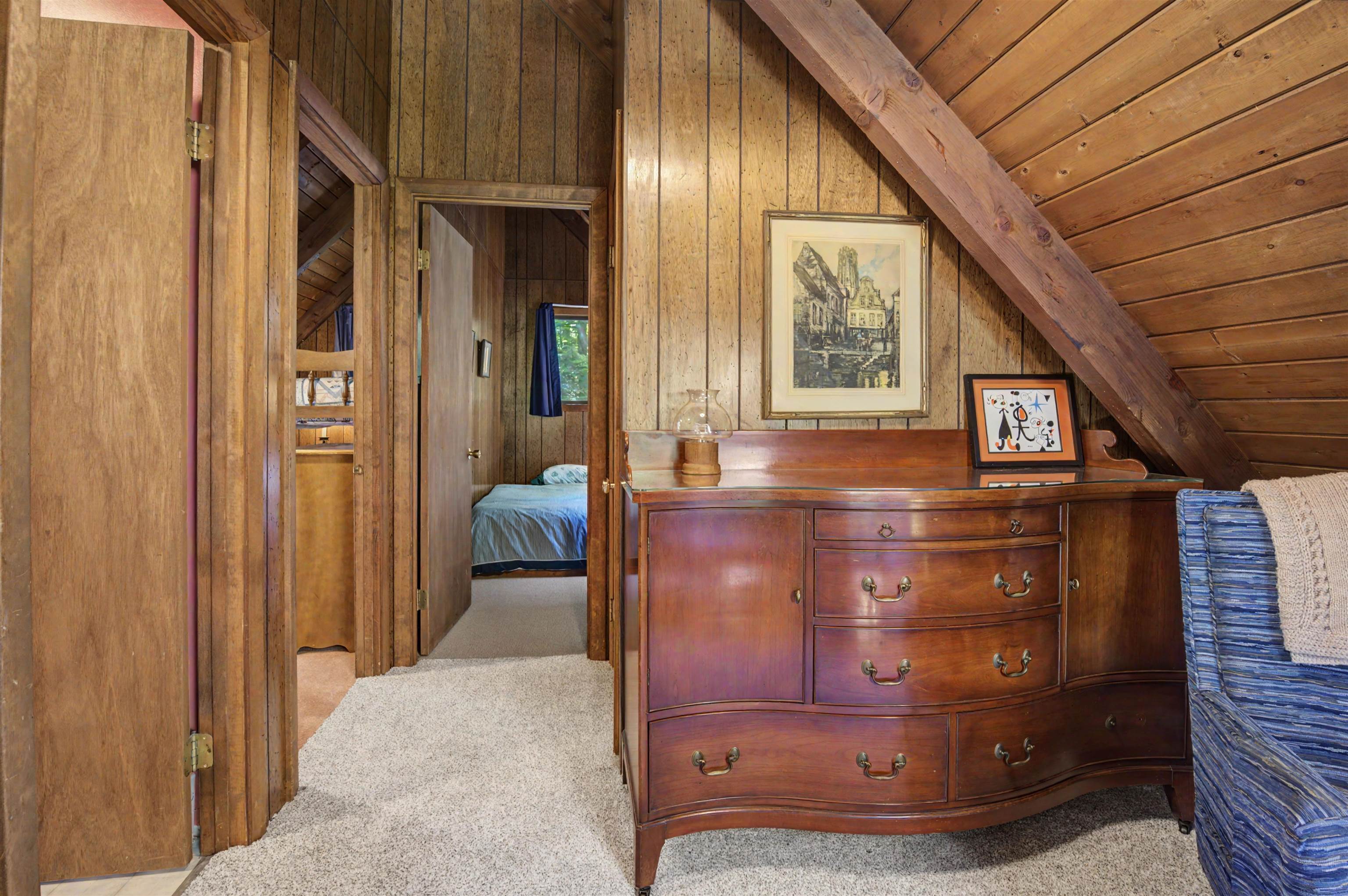 Furnished attic room with sloped wood ceilings, a curved dresser, and a bedroom visible through the doorway.