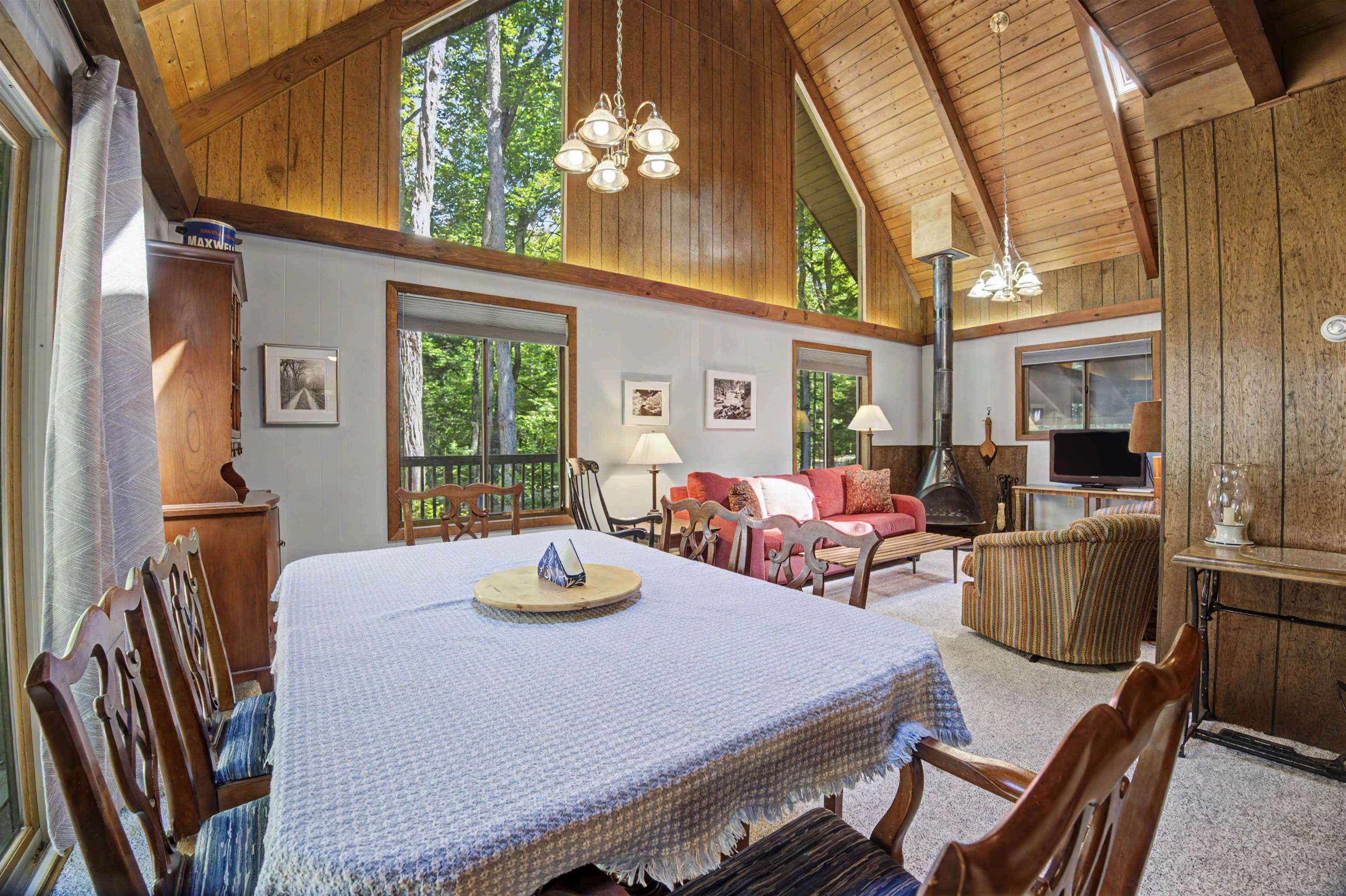 Open-plan living and dining room with vaulted wood ceiling, red sofa, and large forest-facing windows.