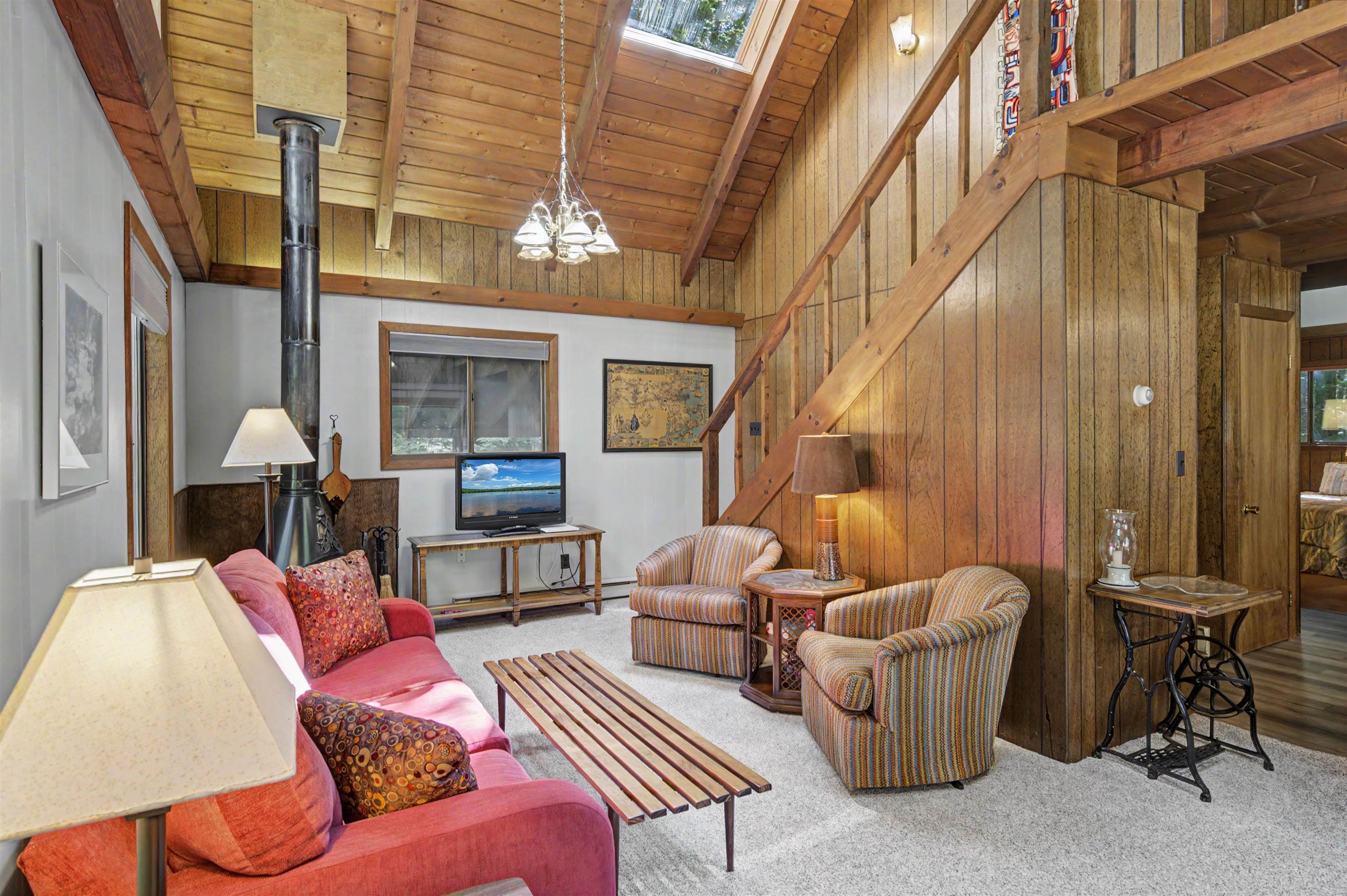 Living room with a vaulted wood ceiling, skylight, wood stove, red sofa, and staircase to a loft.