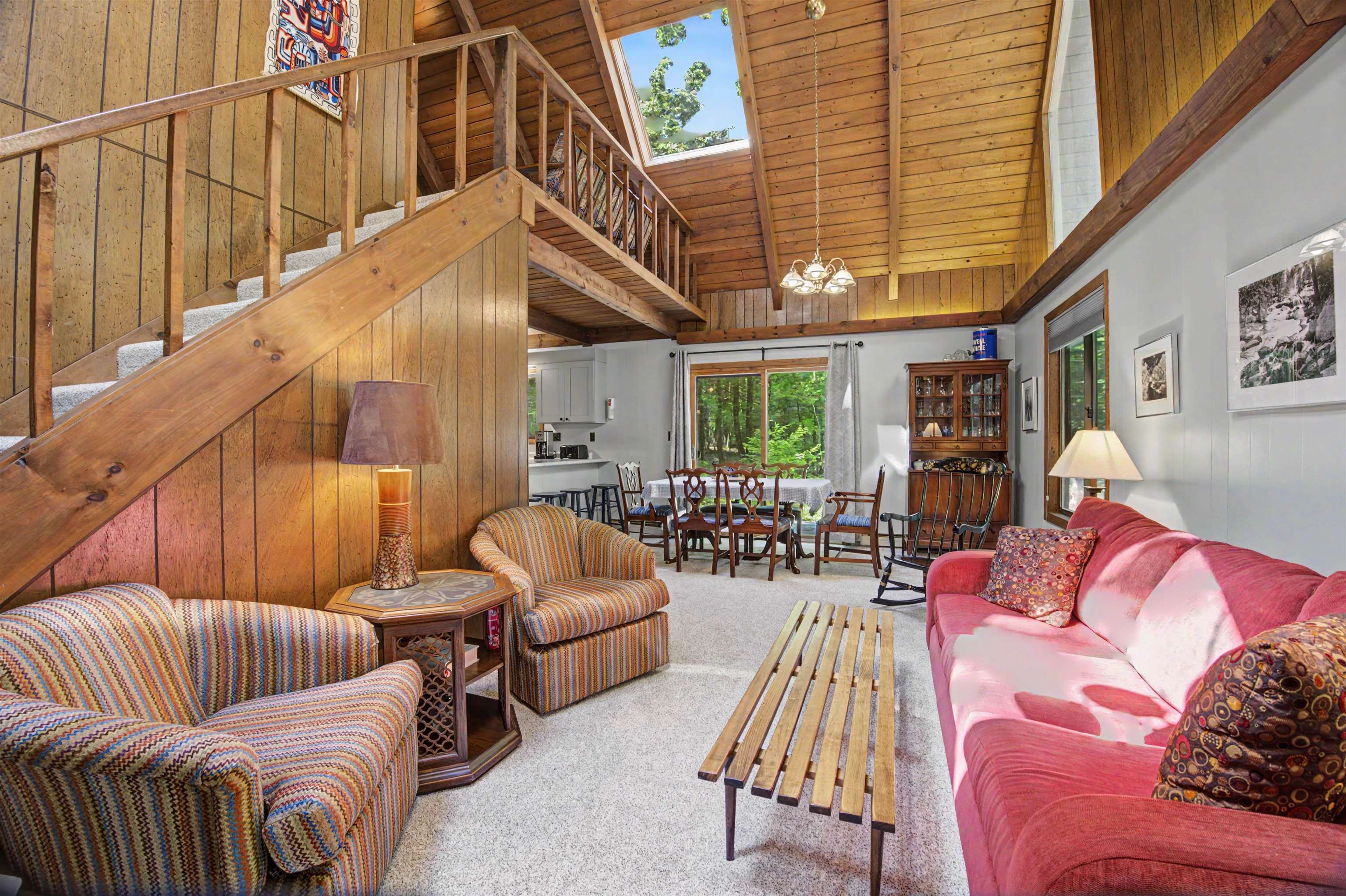 Living room with a vaulted wood ceiling, loft staircase, skylight, patterned armchairs, and pink sofa.