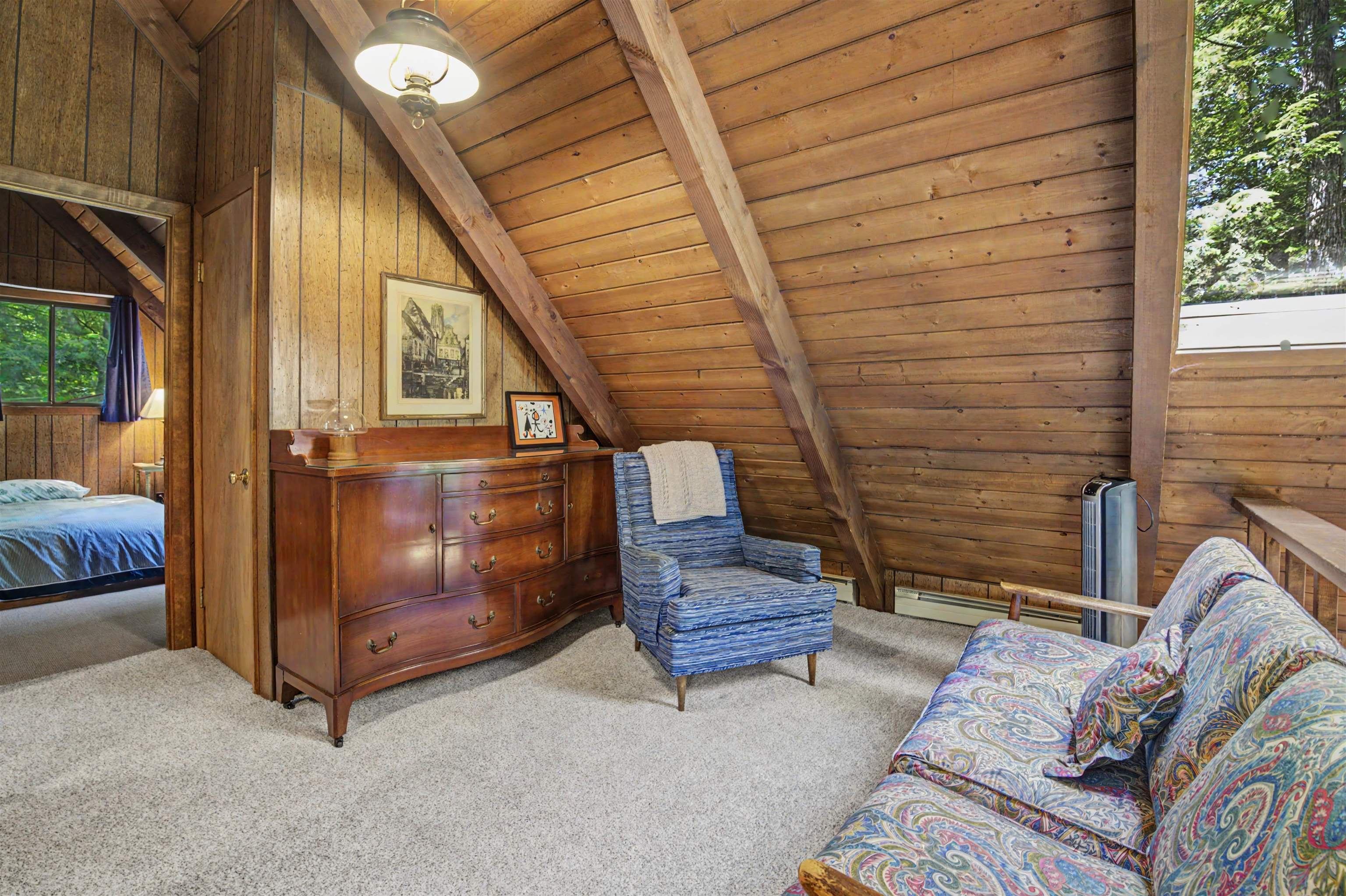 Upper-level sitting area with a sloped wood ceiling, patterned sofa, blue armchair, and doorway to a bedroom.