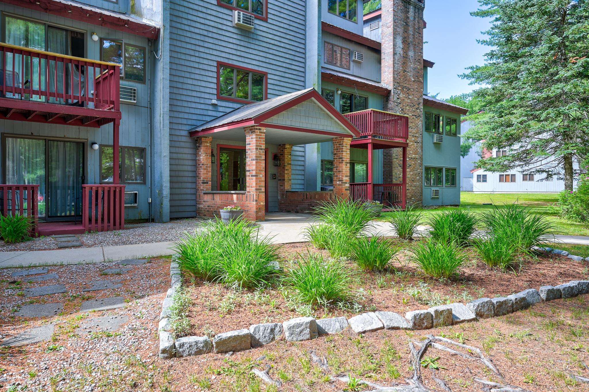 Apartment building entrance with brick columns, red balconies, and a landscaped garden.