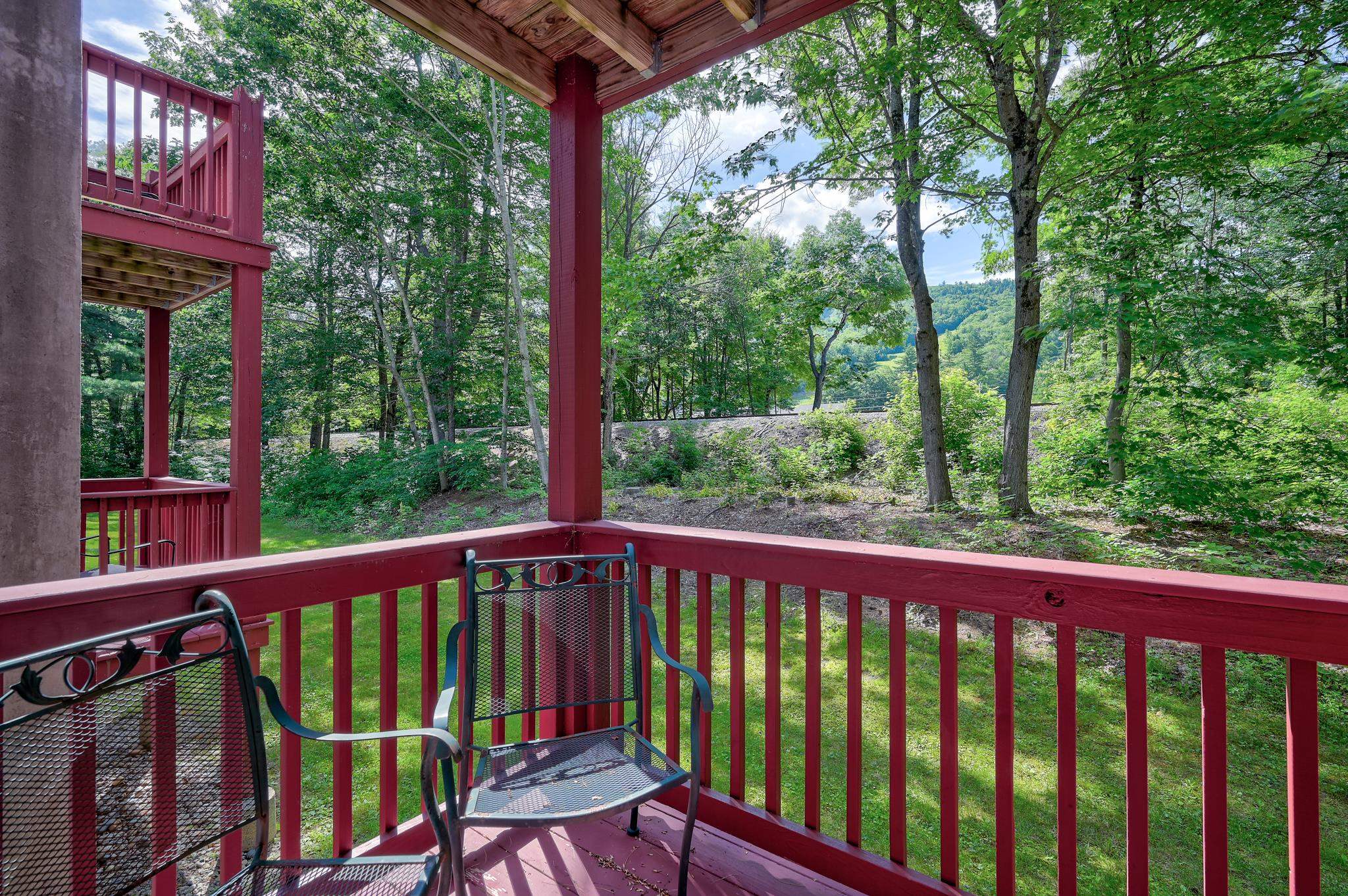 Covered deck with red railings and metal chairs overlooking a wooded lawn and distant hillside.
