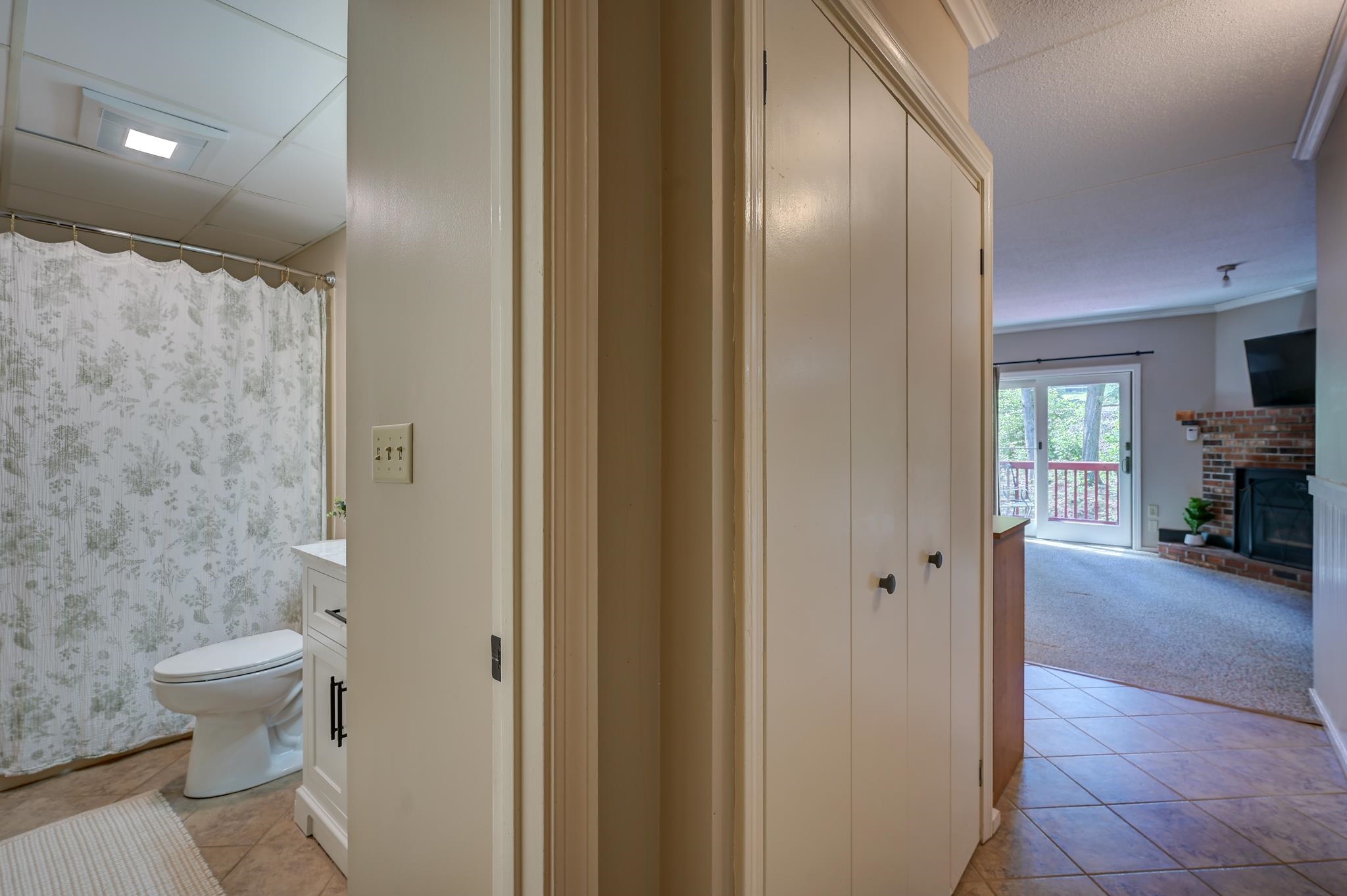 Bathroom with patterned shower curtain and built-in storage, looking toward a living room with a brick fireplace.