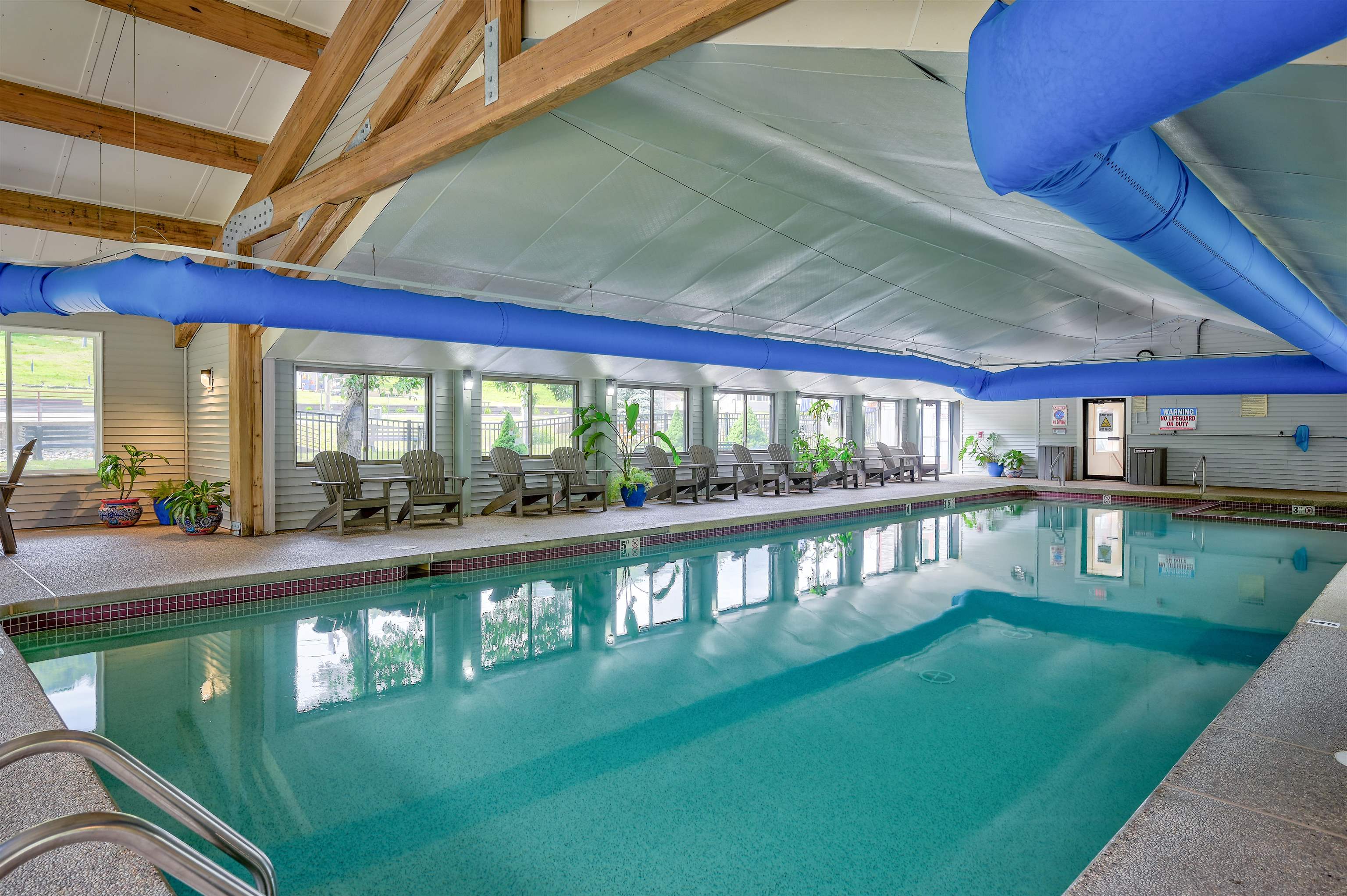 Indoor swimming pool beneath exposed timber beams, with blue ducts and chairs along the windowed deck.
