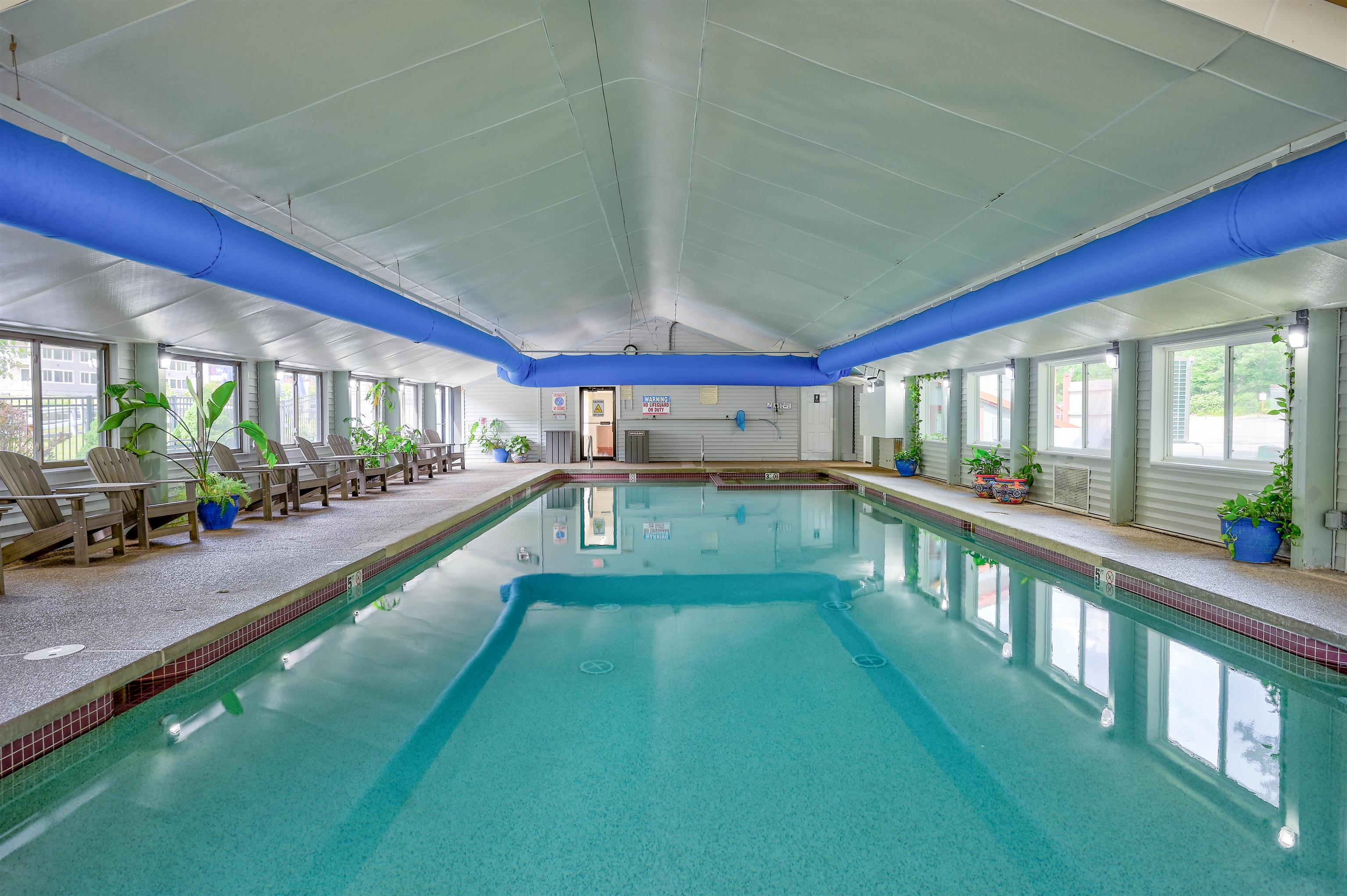 Indoor pool with blue ceiling ducts, large windows, and wooden lounge chairs along the deck.