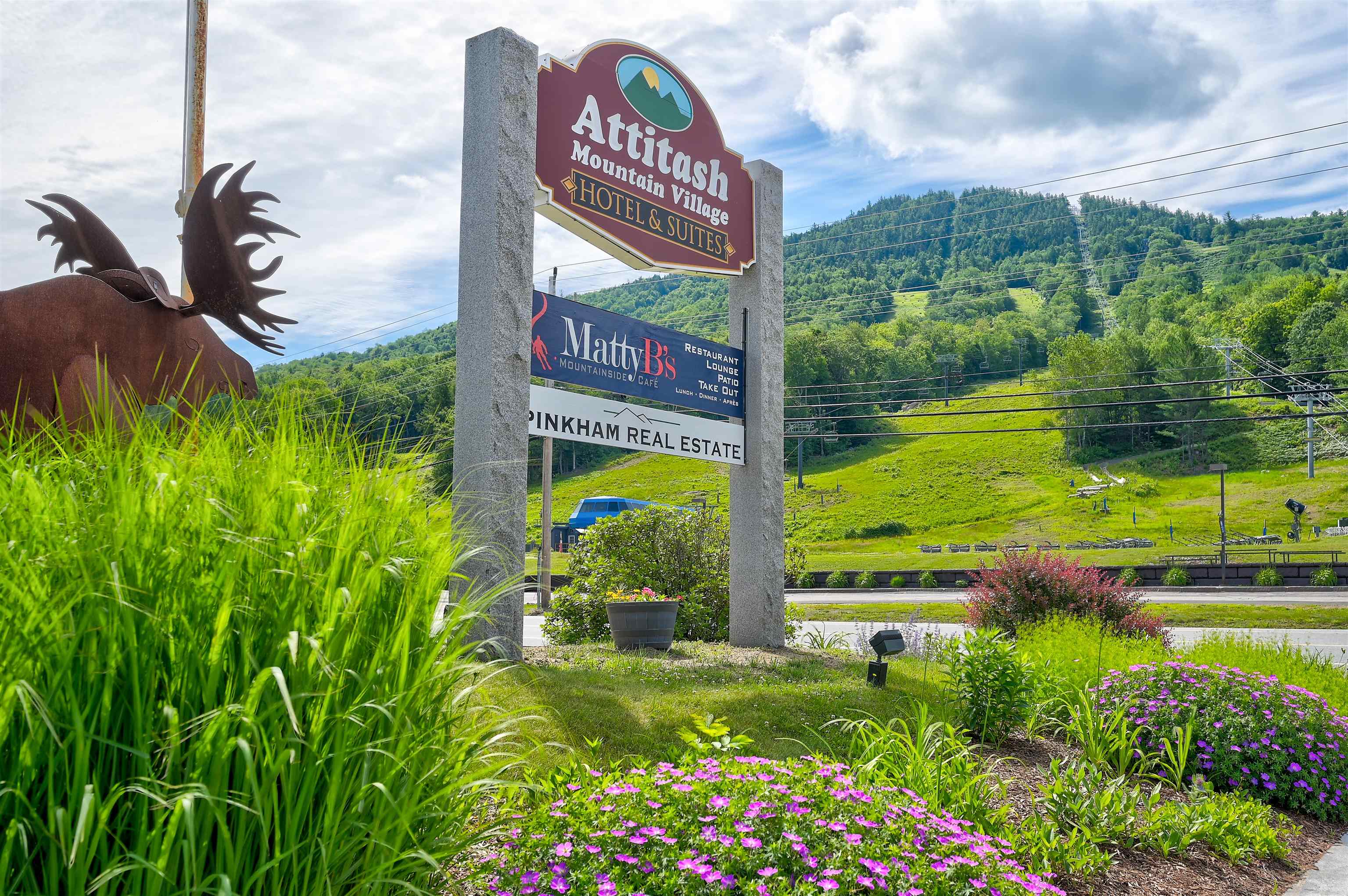 Attitash Mountain Village sign beside a green ski slope, with a moose sculpture and colorful flower beds.