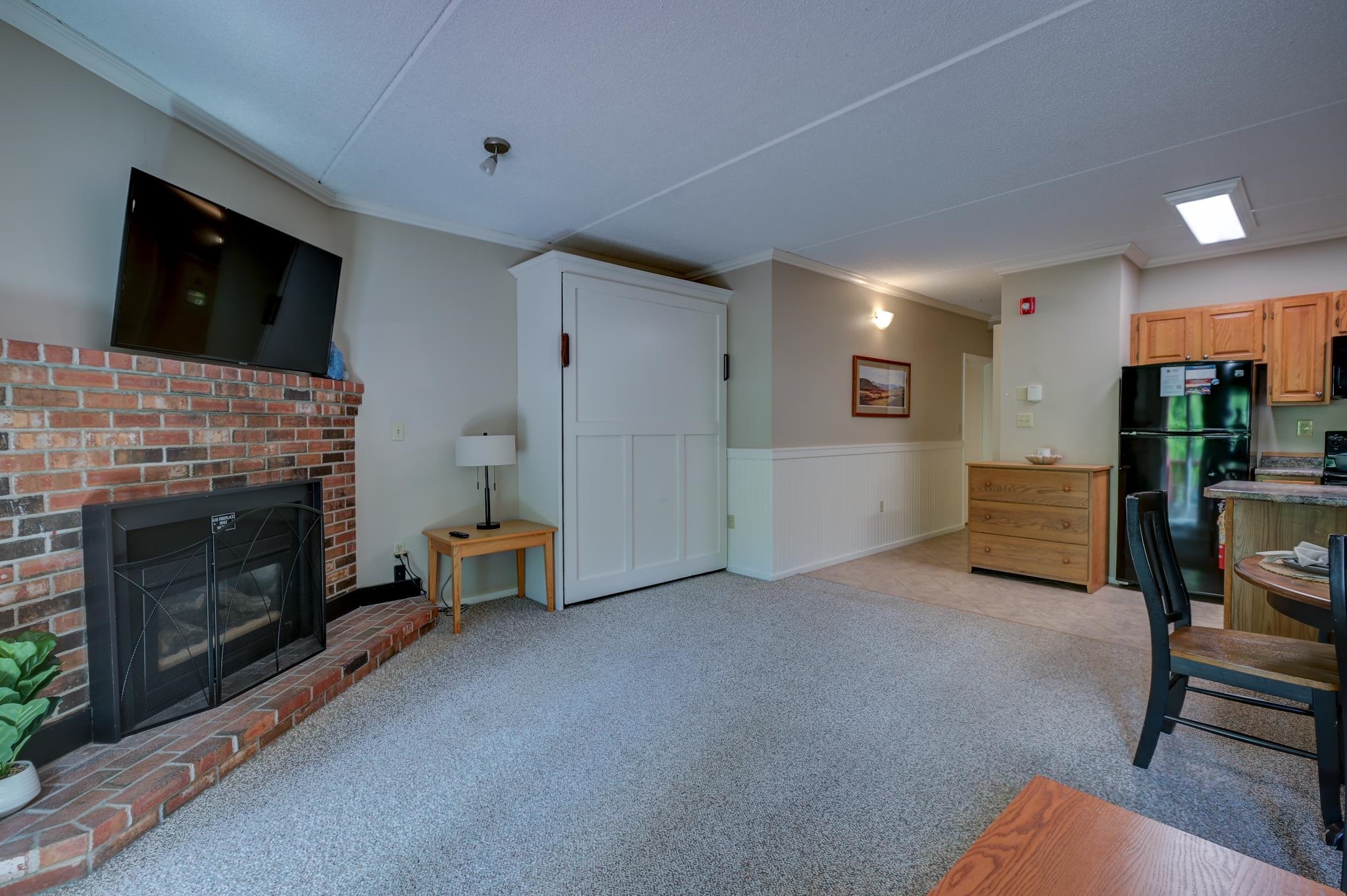 Living room with a brick fireplace, mounted TV, white Murphy bed, and adjoining kitchen.