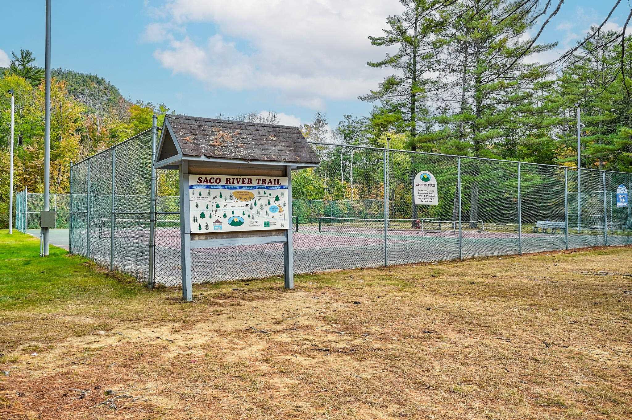 Fenced tennis courts beside a covered Saco River Trail map sign, with autumn trees in the background.