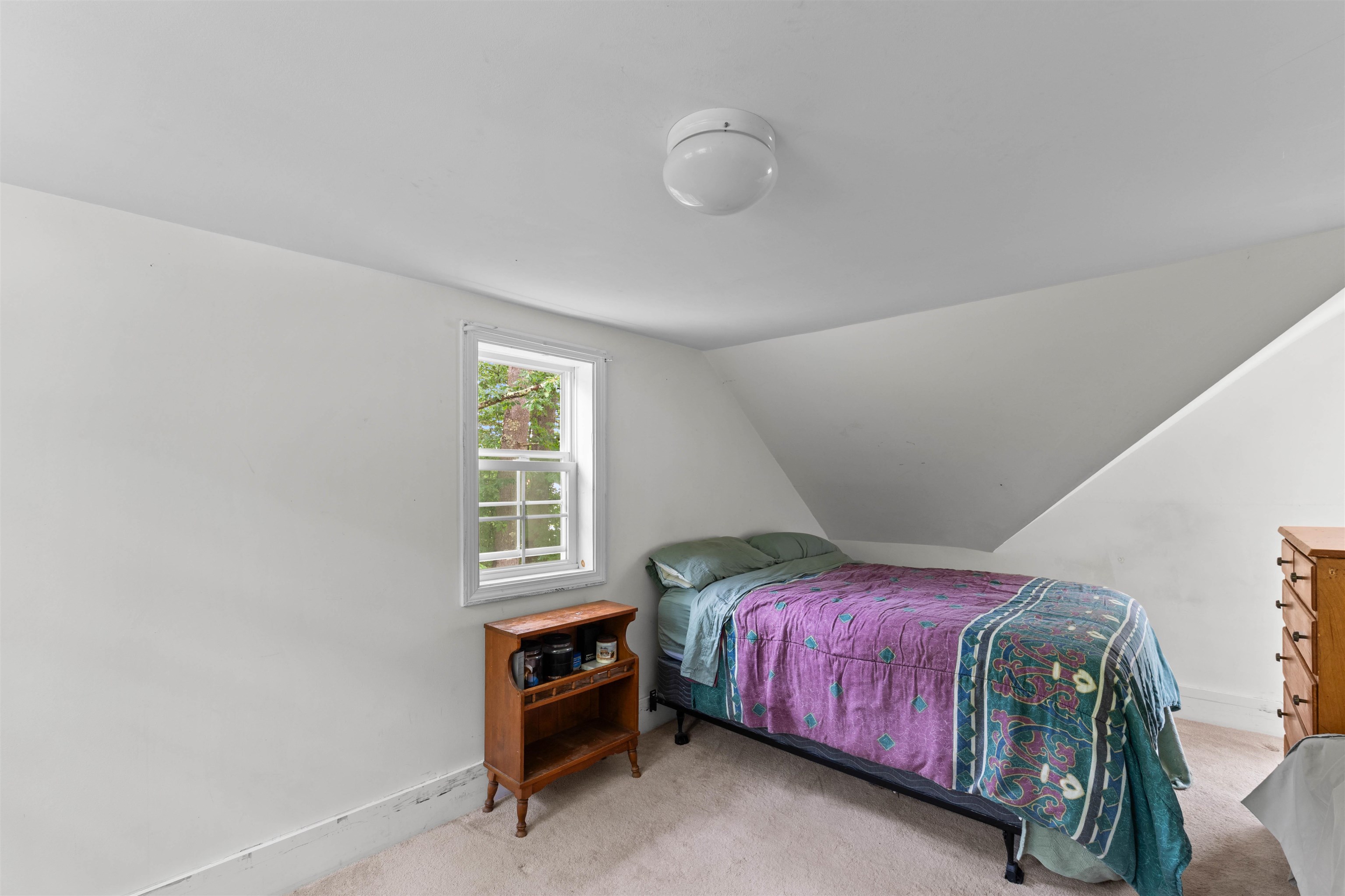 Attic bedroom with a purple and teal quilt, sloped ceiling, and window overlooking trees.
