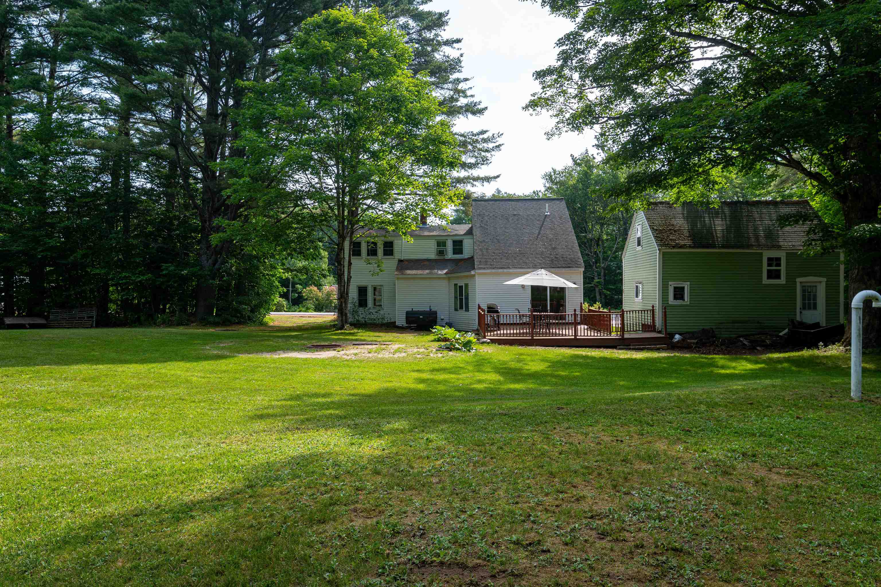 House with a rear deck and umbrella beside a green outbuilding, surrounded by trees and lawn.