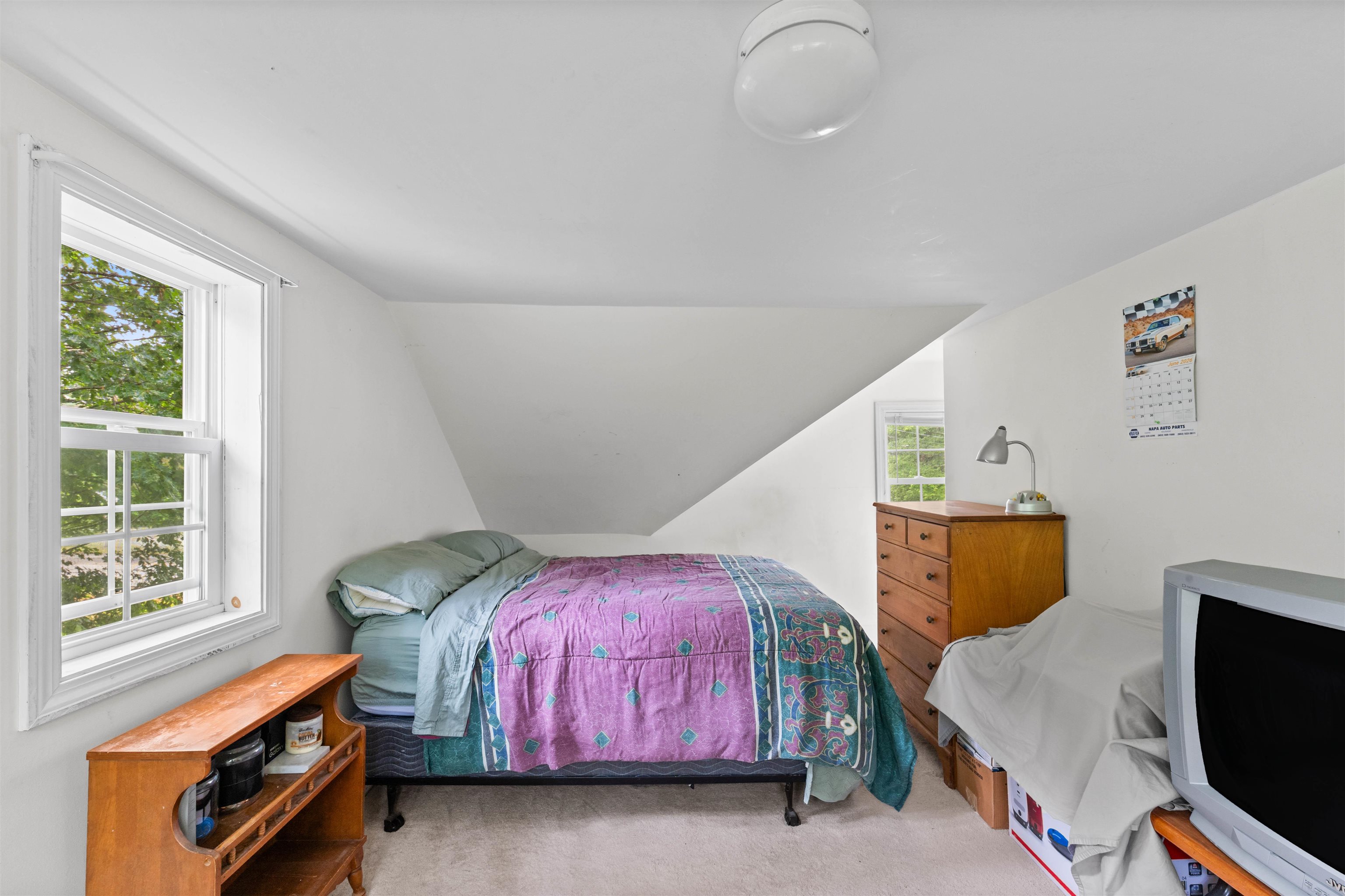Upstairs bedroom with a purple quilted bed beneath a sloped ceiling and a large window overlooking trees.