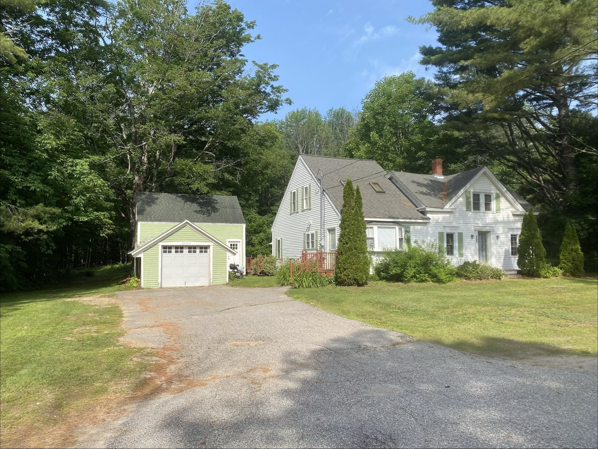White multi-gabled house with green shutters, a detached garage, and a wooded setting.