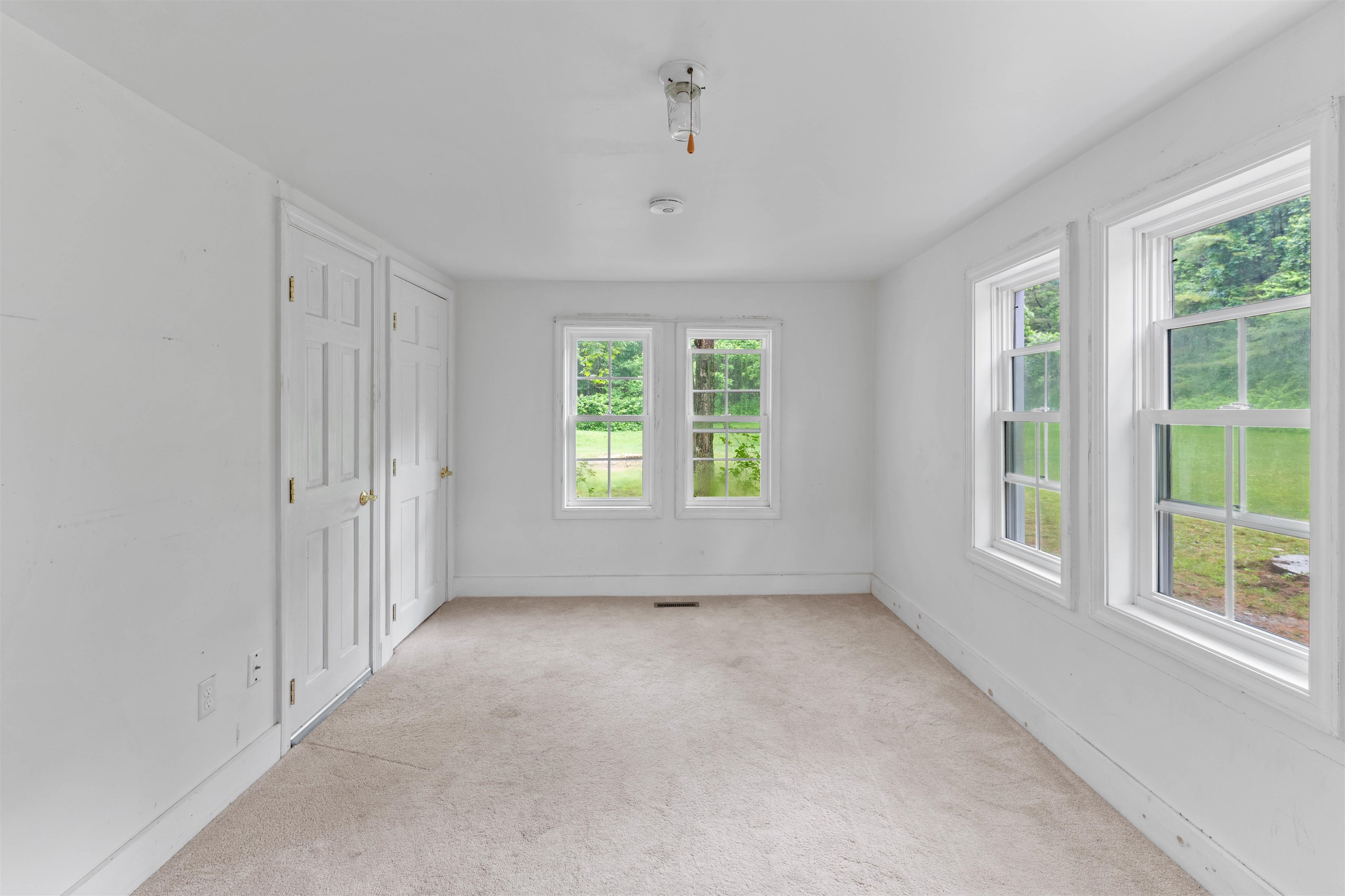 Vacant carpeted room with white walls, paneled doors, and four windows overlooking a green lawn.