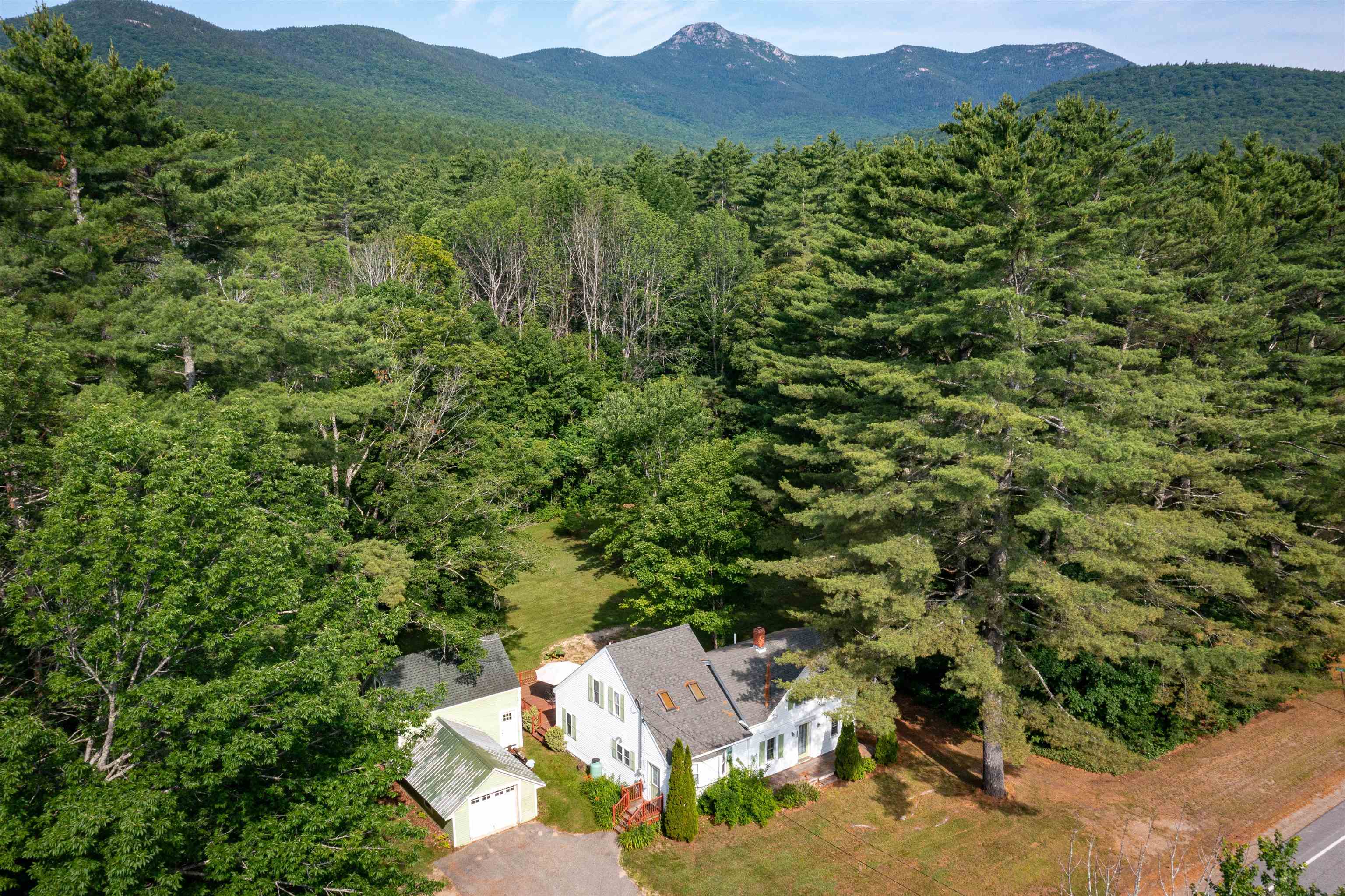 Aerial view of a white house and garage amid dense trees, with mountains beyond.