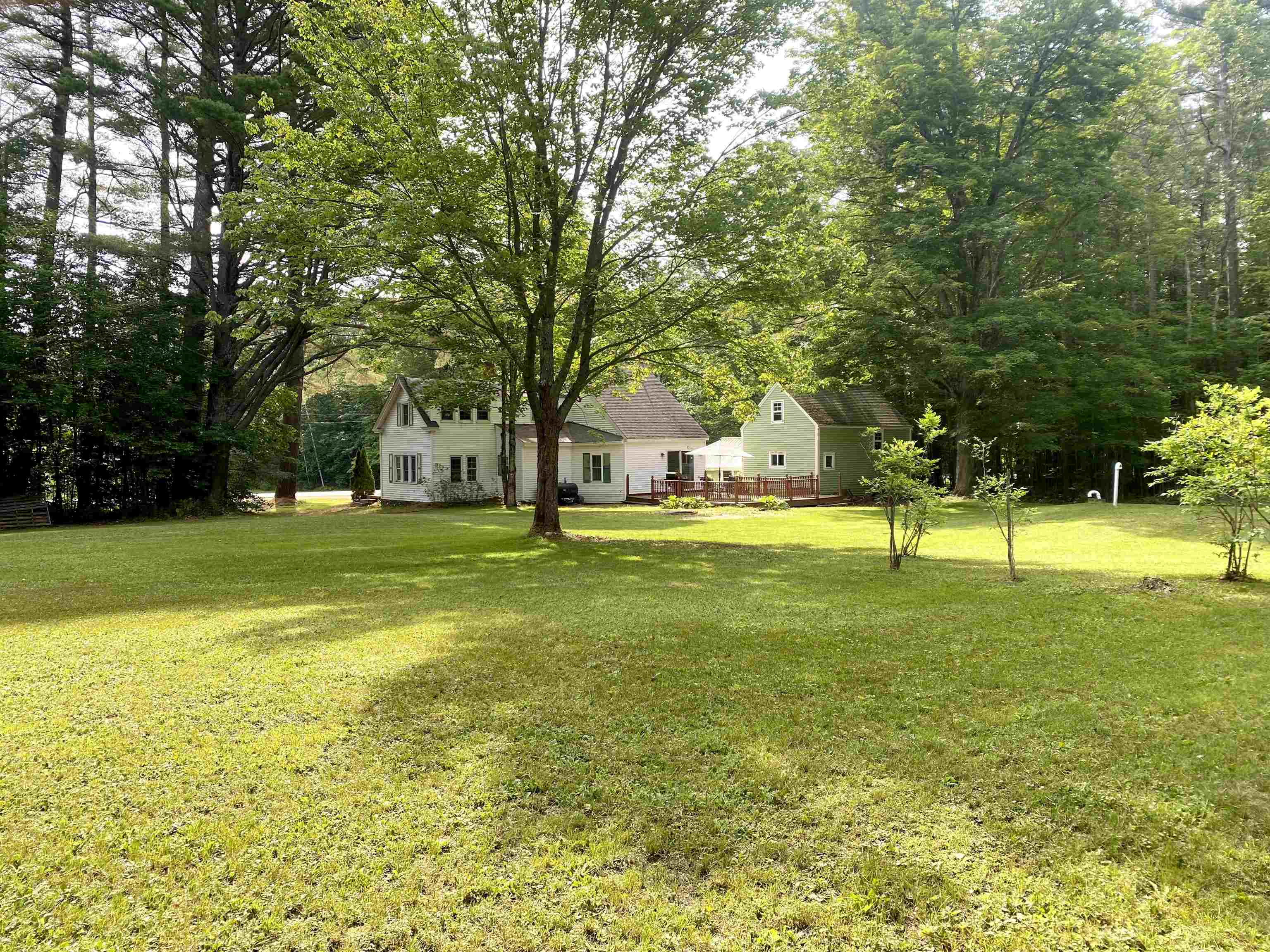 Light-colored house and outbuilding nestled among trees beside a broad lawn and deck.