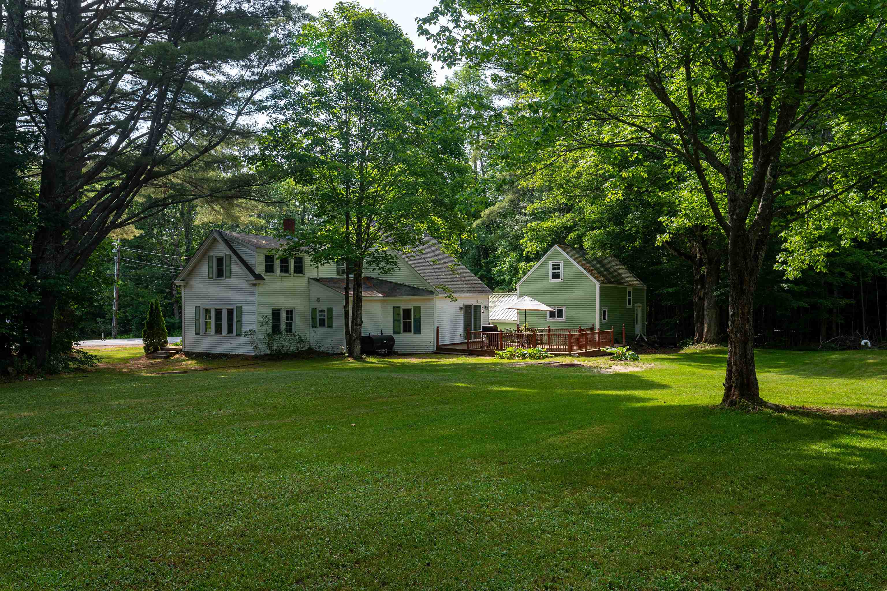 Light-colored house and green outbuilding with a deck, framed by mature trees and a broad lawn.