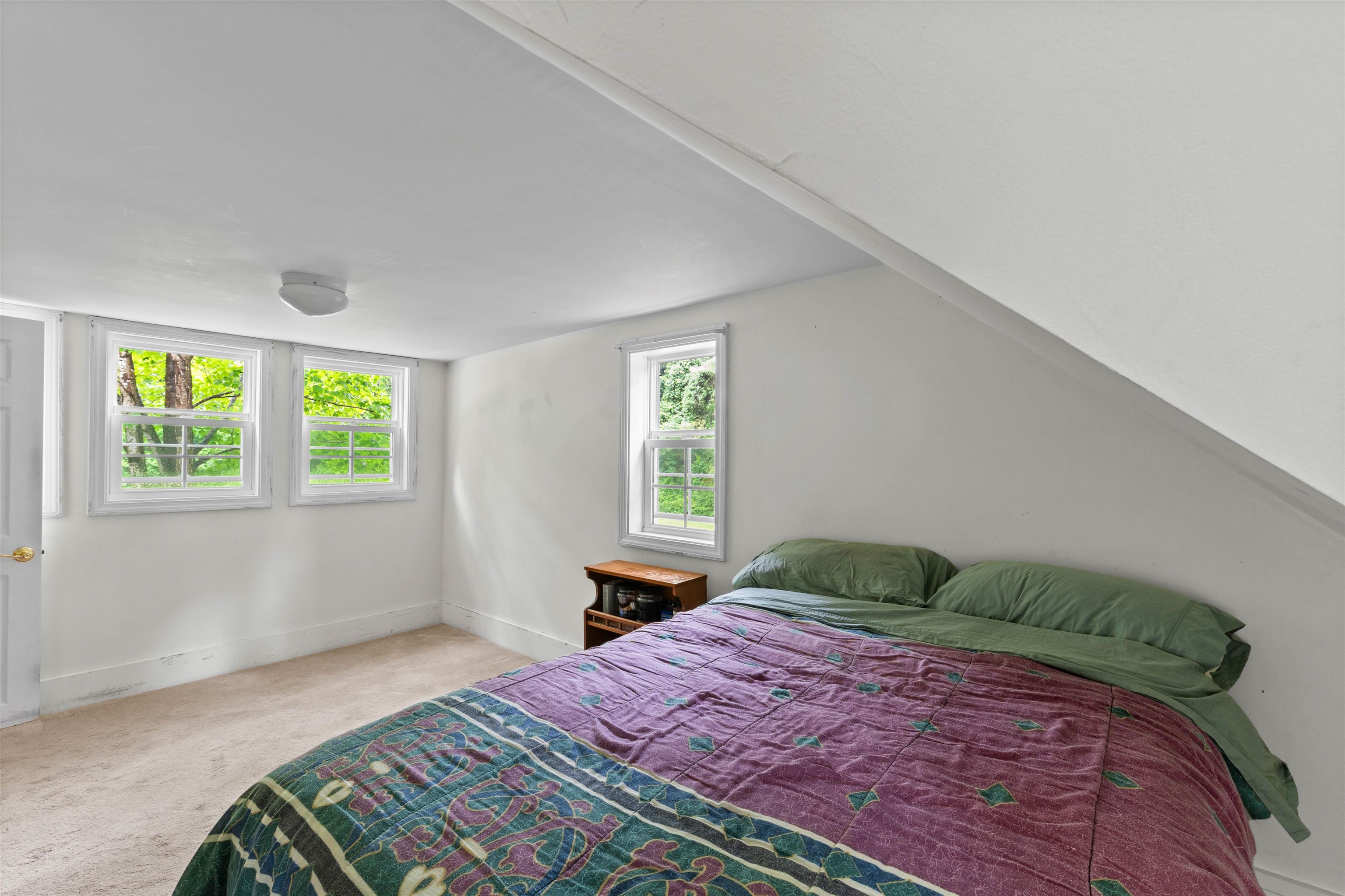 Bedroom with a sloped ceiling, a purple patterned quilt, beige carpet, and several windows.