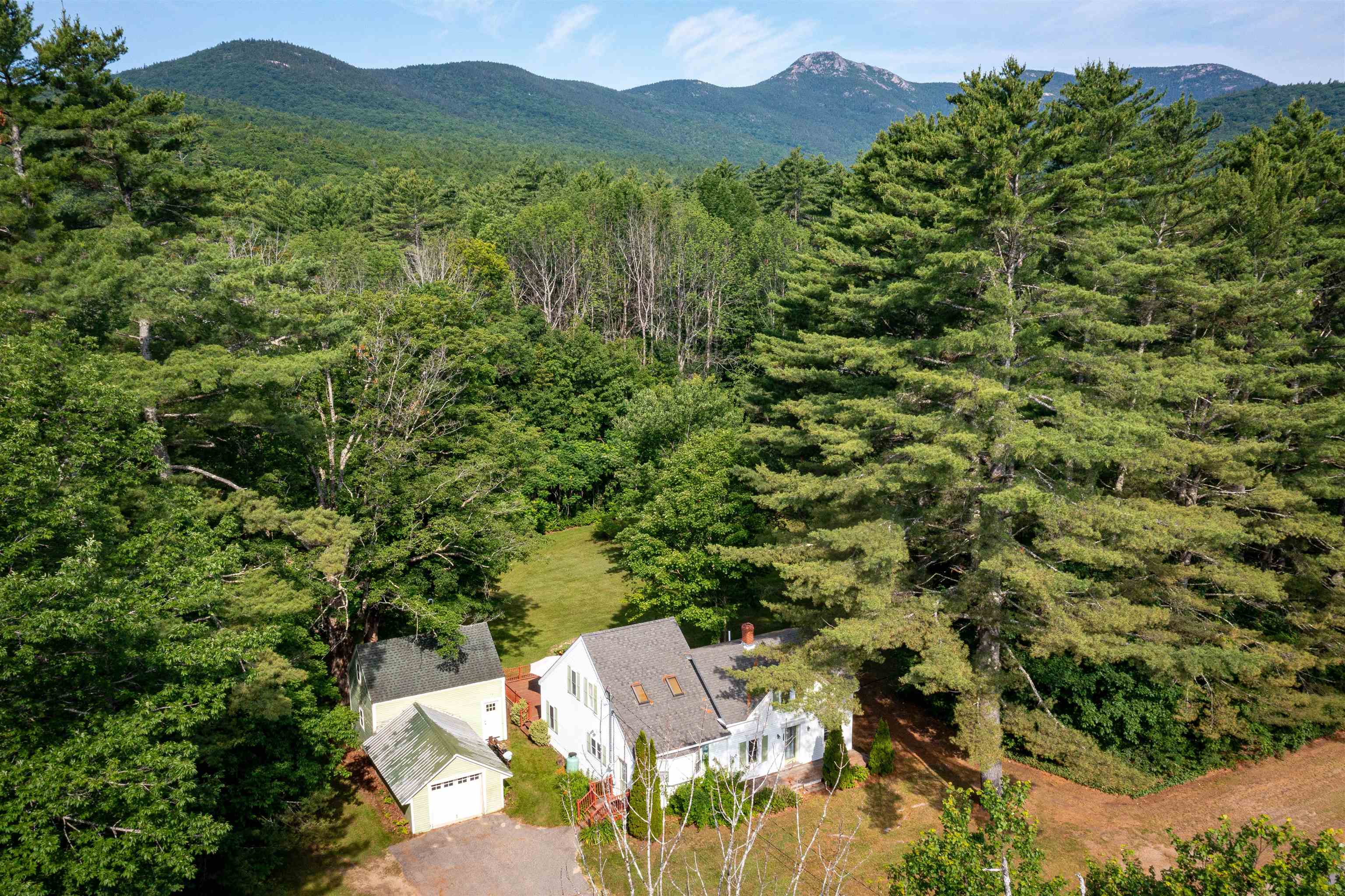Aerial view of a white house and detached garage amid forest, with mountains beyond.