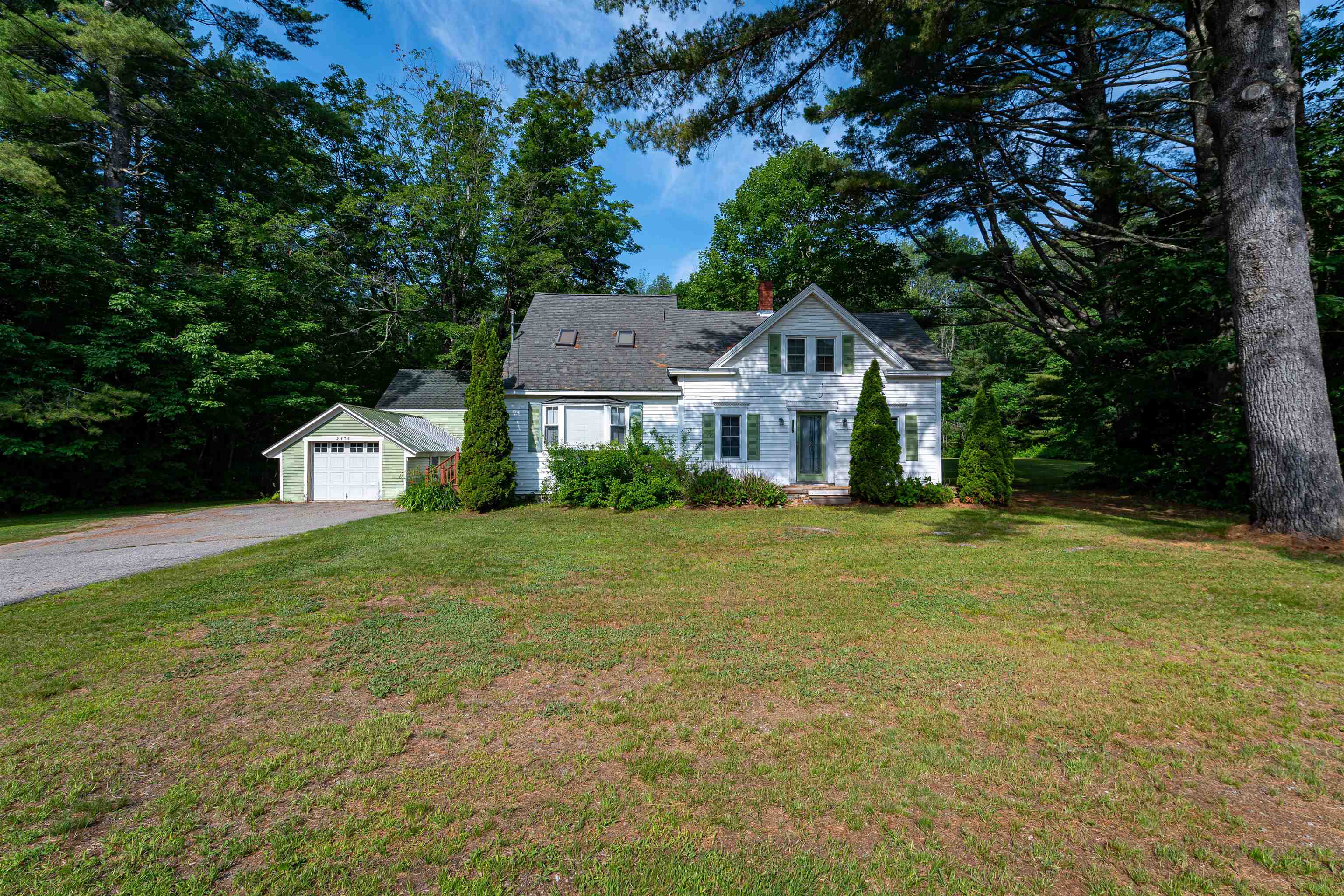 White house with green shutters, detached garage, and gravel driveway beneath mature trees.