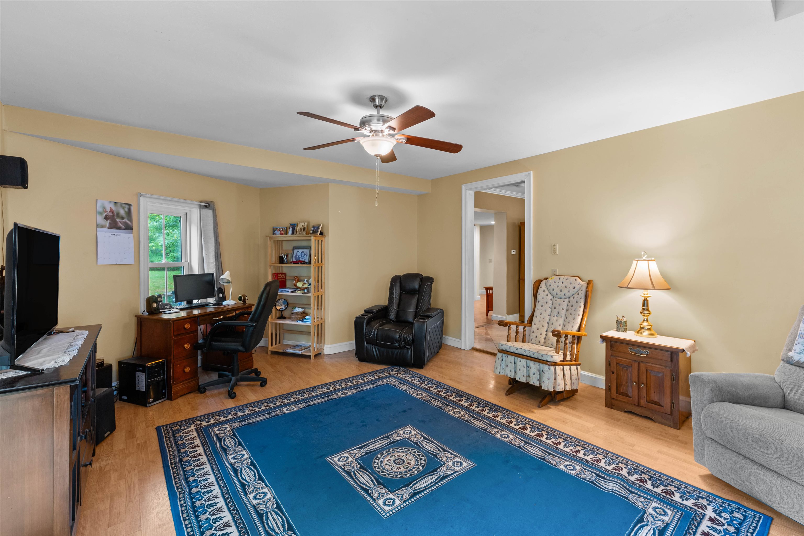 Furnished living room with a blue patterned rug, desk by the window, and ceiling fan.