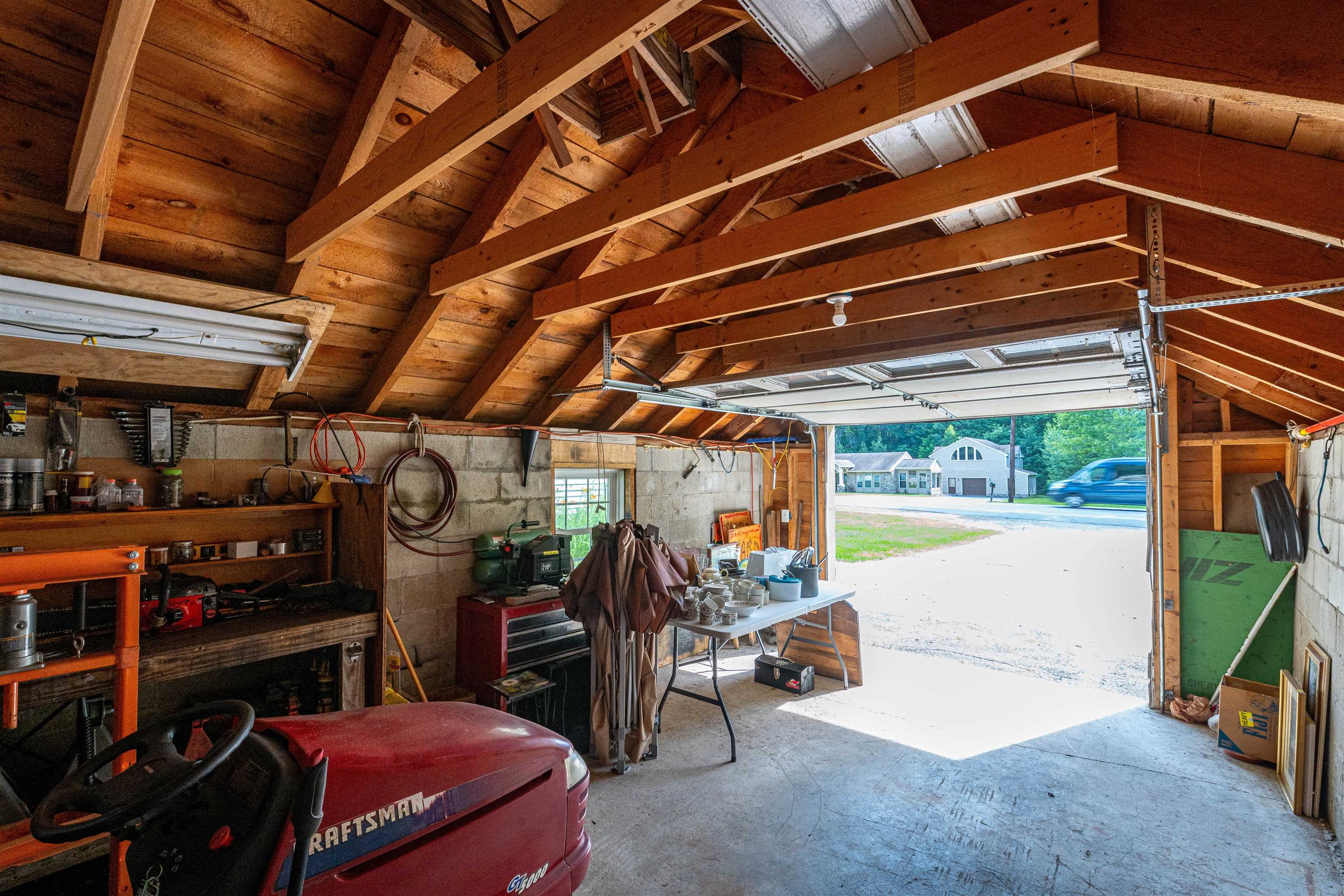 Garage with exposed timber rafters, workshop storage, and a red Craftsman riding mower by the open door.