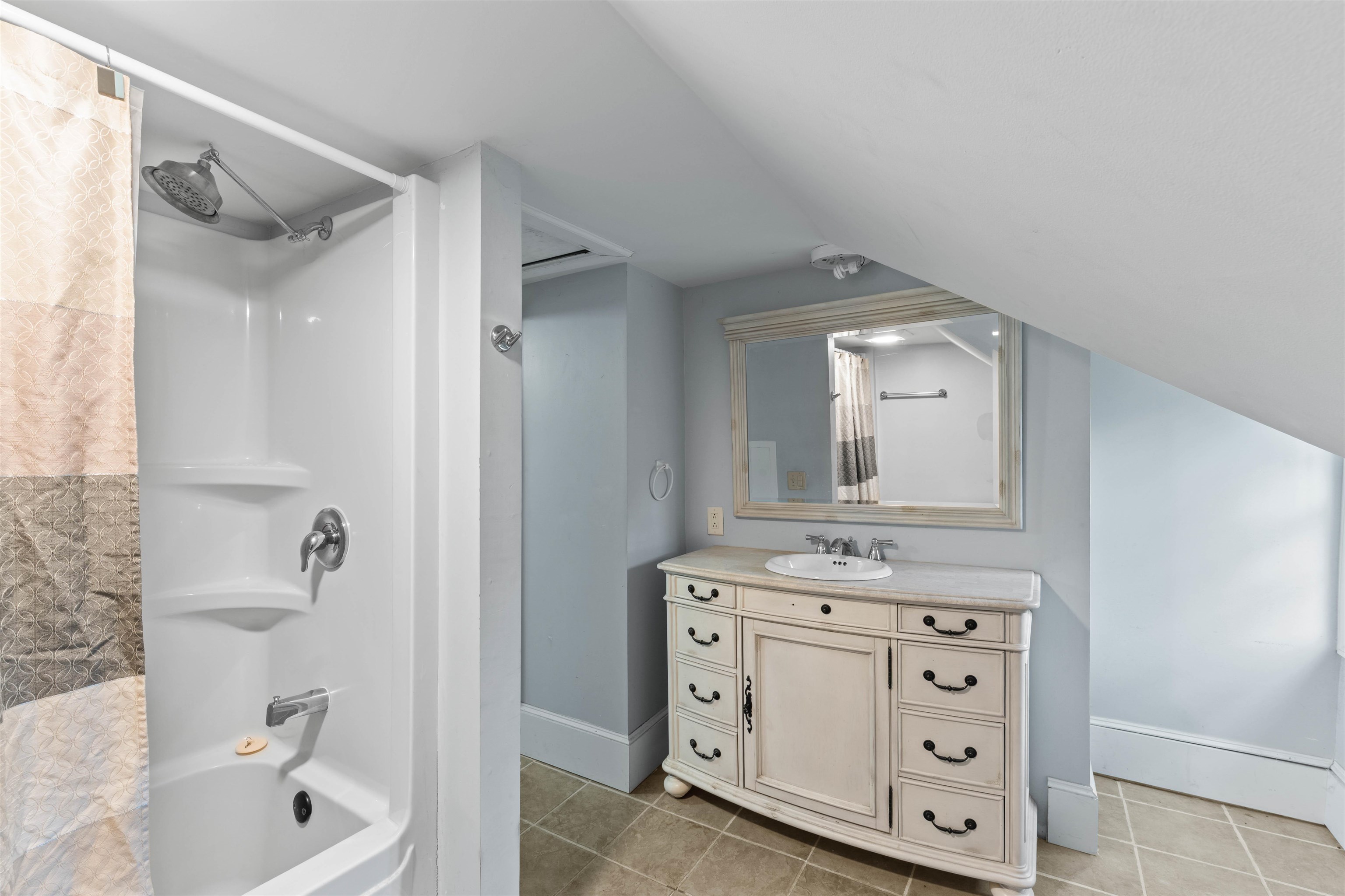 Attic bathroom with a white shower-tub and patterned curtain beside a cream vanity and framed mirror.