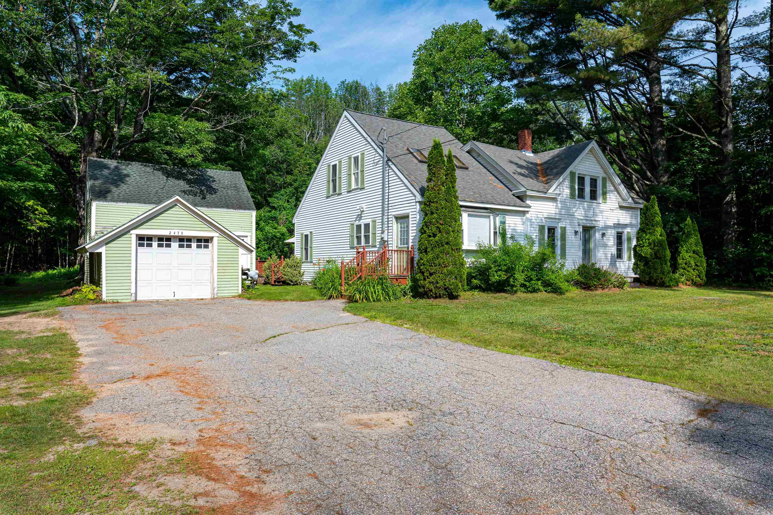 White multi-gabled house with a detached garage and broad driveway, surrounded by mature trees.