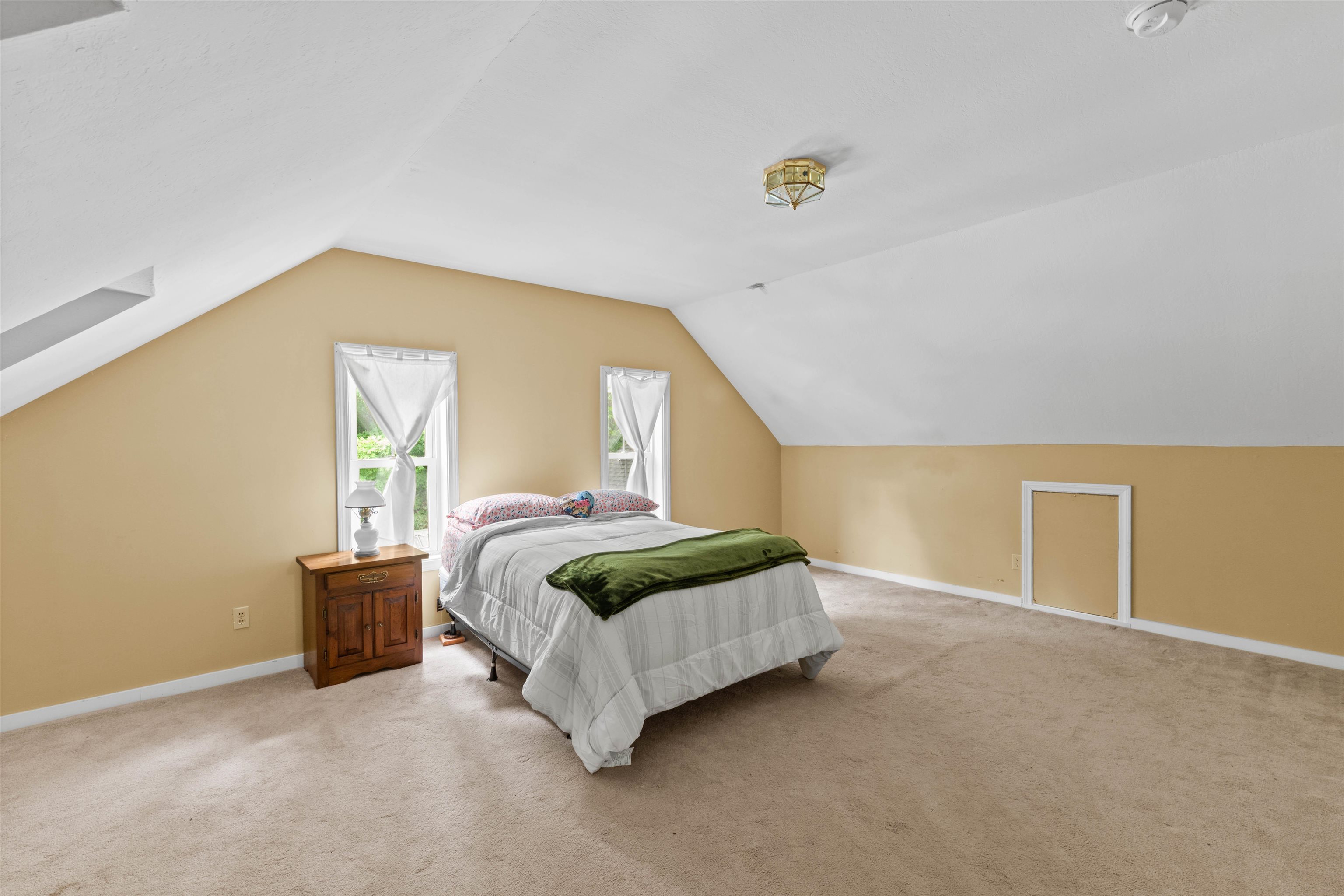Furnished bedroom with a bed beneath a sloped ceiling and two curtained windows.