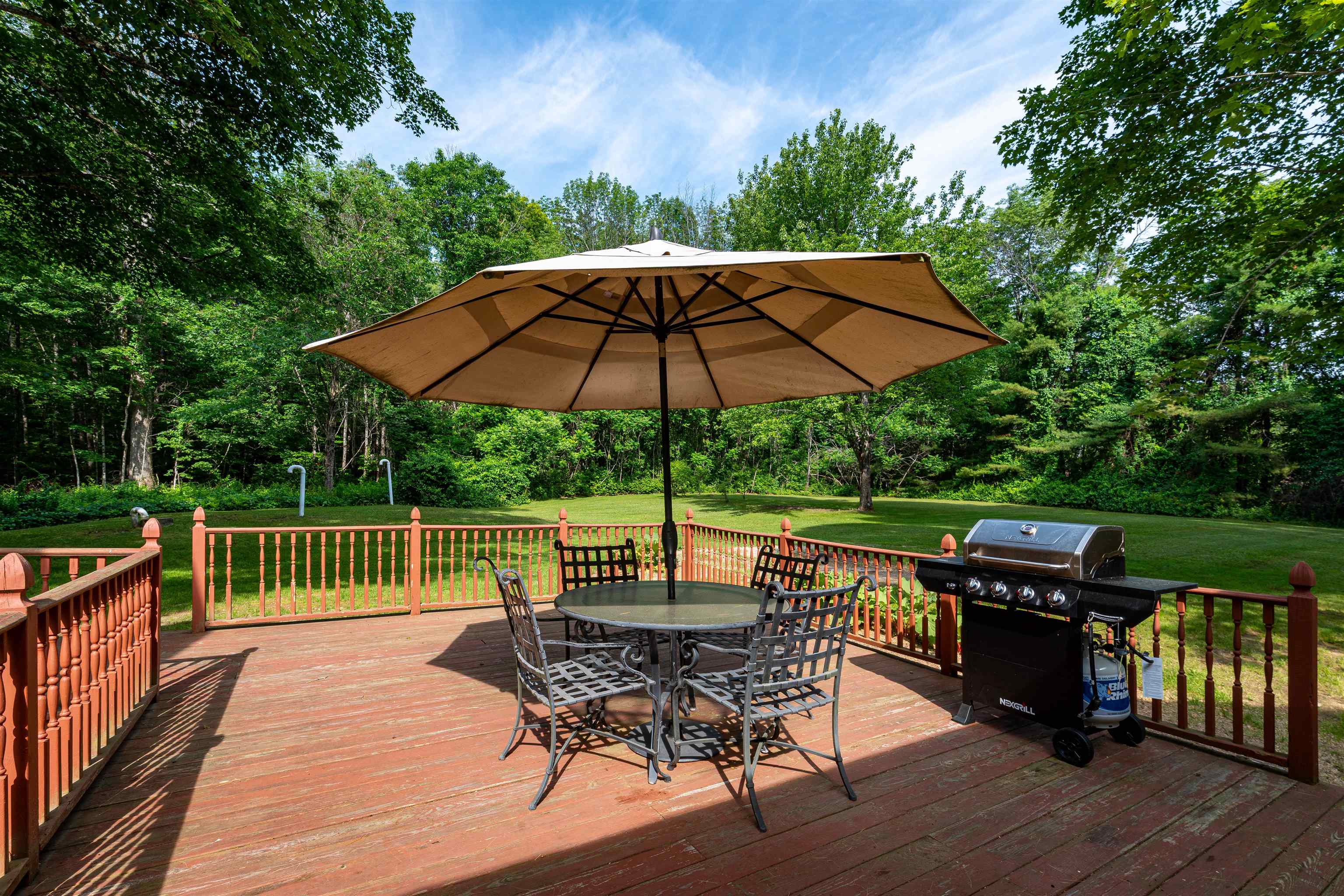 Wooden deck with patio dining set under a large umbrella and a gas grill beside a wooded lawn.