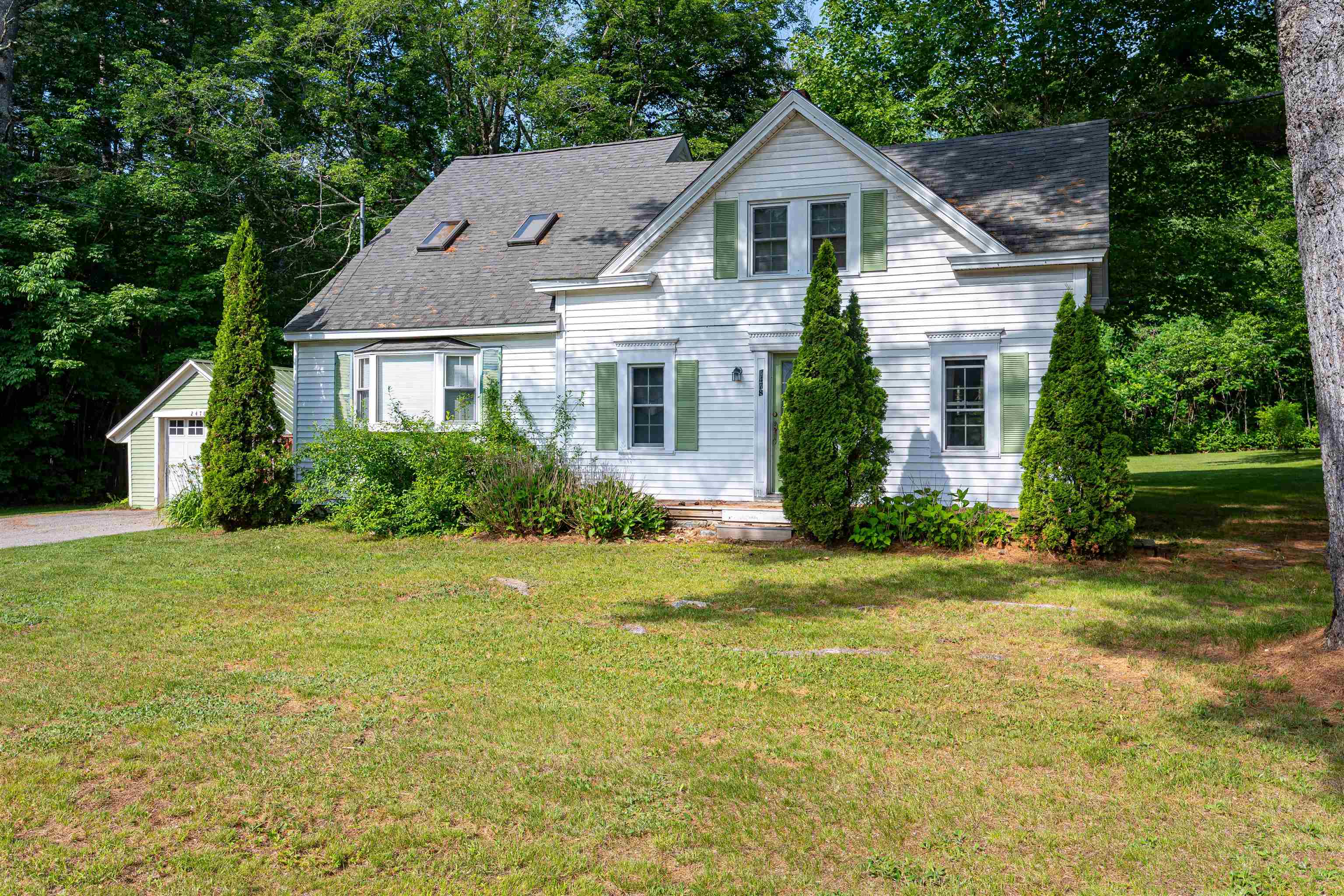White two-story house with green shutters and attached garage, surrounded by trees and a broad lawn.